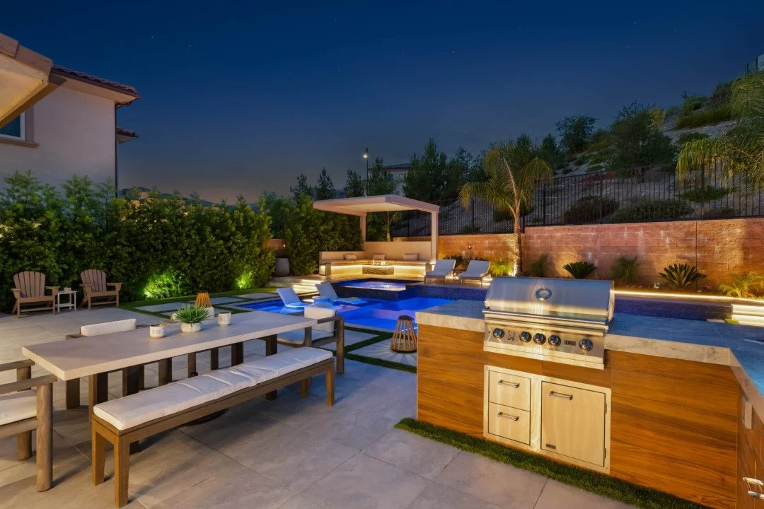 Nighttime outdoor backyard with illuminated swimming pool, lounge chairs, seating area with cushions, and a built-in outdoor grill.
