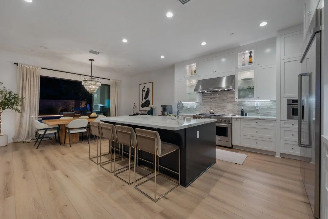 Modern open-concept kitchen with white cabinets, stainless steel appliances, a large black and white island with seating, hardwood floors, and a dining area with a chandelier and large window with curtains.