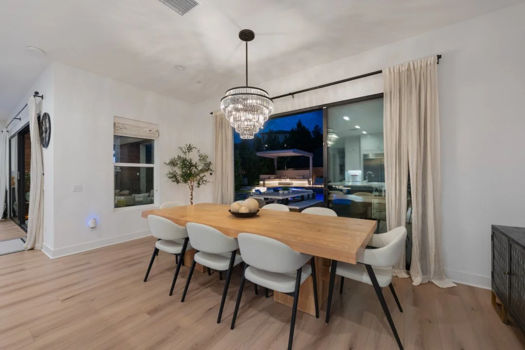 A dining room with a wooden table, six white chairs, a chandelier, a window, and glass sliding doors leading to an outdoor patio at dusk.