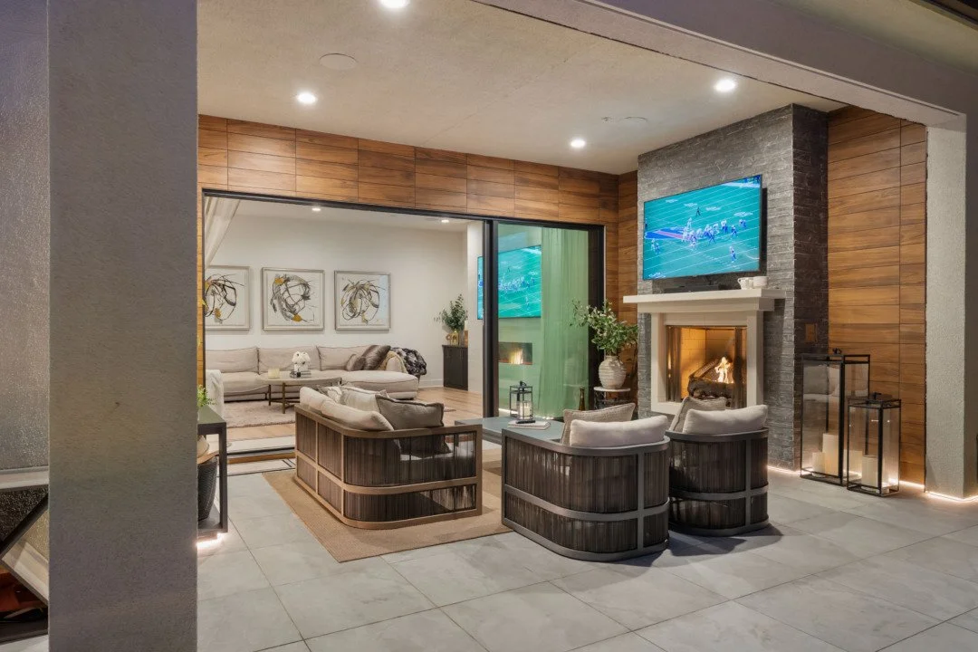 A cozy living room with a fireplace, wall-mounted TV, and seating area, adjacent to another sitting space separated by a sliding glass door, decorated with artwork and plants.
