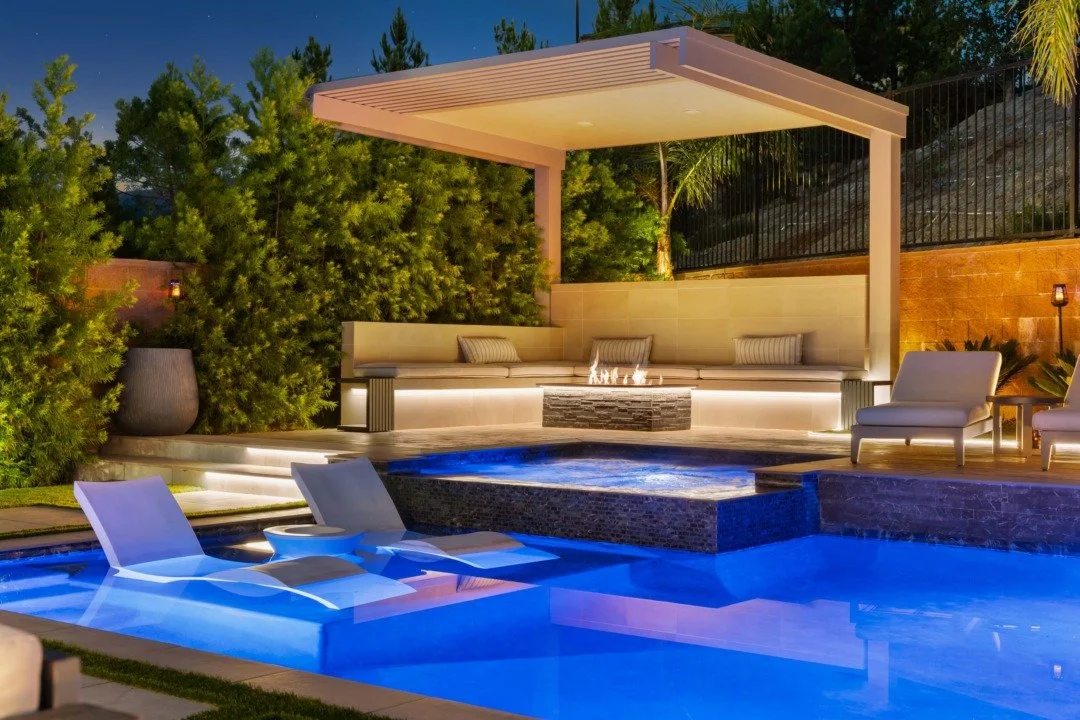 Night view of a modern backyard with a lit swimming pool, outdoor lounge area with a shaded pergola, and lush greenery.