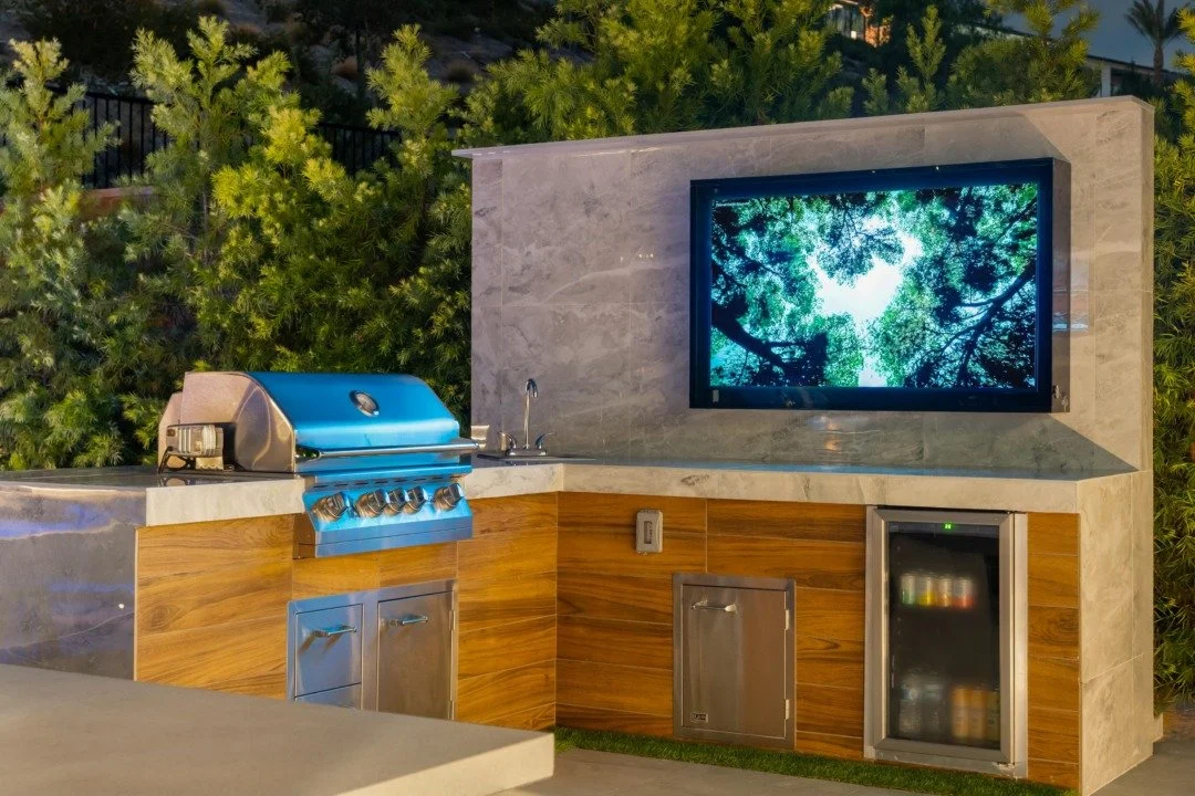Outdoor kitchen with a grill, a sink, and a flat-screen TV mounted on a stone wall, with green foliage behind.