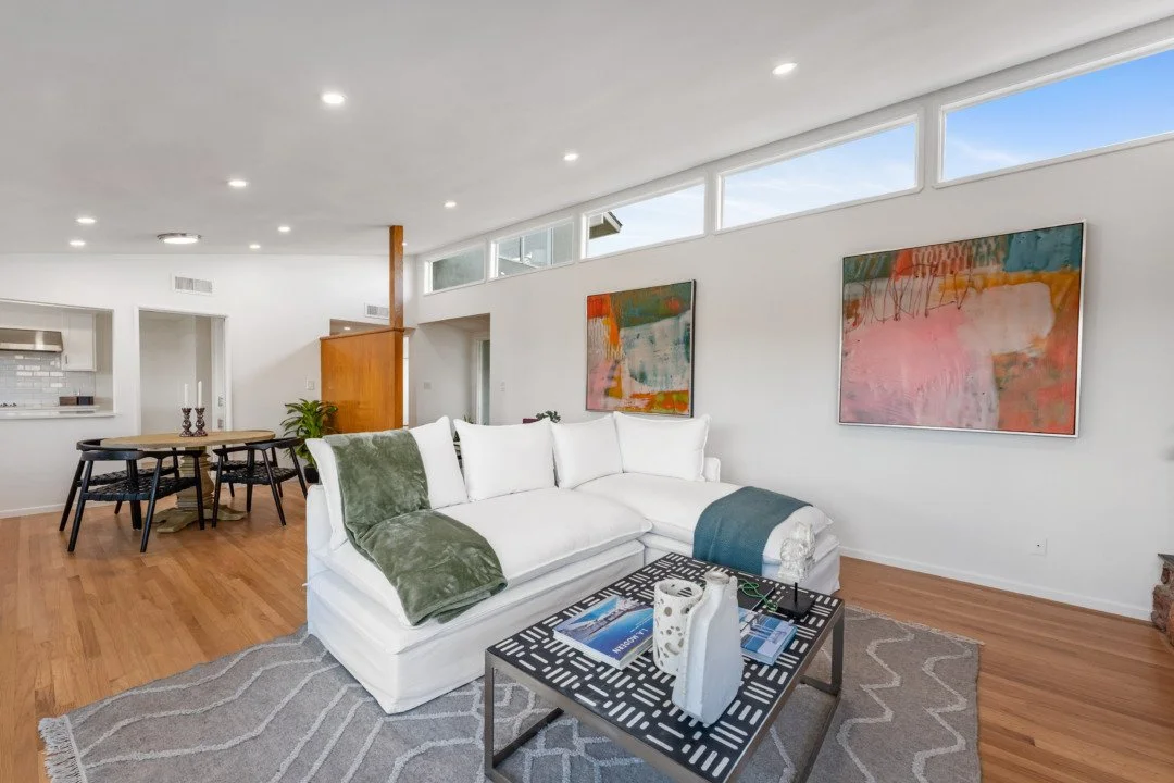 Living room with white wall, modern abstract paintings, white sofa with green and blue throws, coffee table with magazines and decor, hardwood floors, and large windows near ceiling.