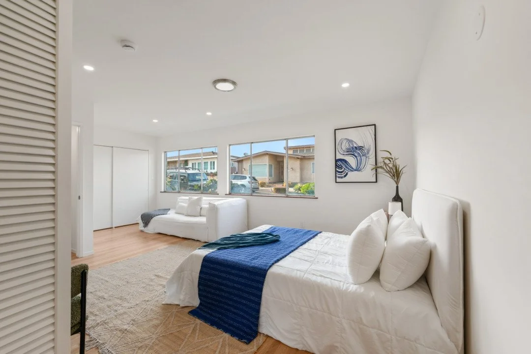 Bright bedroom with white walls, a bed with white bedding and a blue throw, and a white couch by the window with a blanket. There is artwork on the wall and a vase with dried branches. Large windows show parked cars and houses outside.