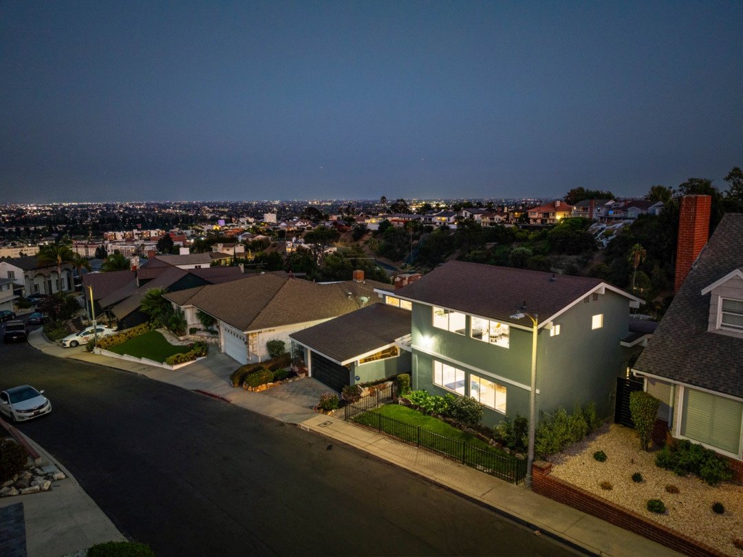 Aerial night view of a suburban neighborhood with lit houses, streets, and city lights in the distance.