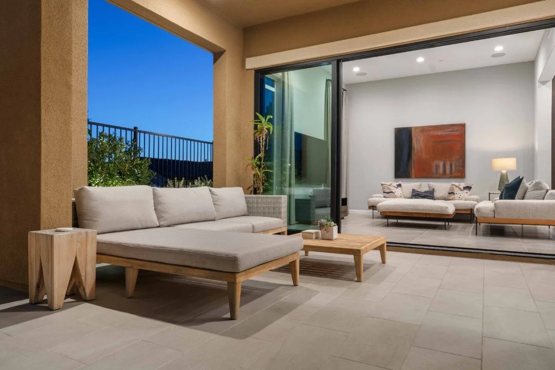 Balcony outdoor seating area with a sectional sofa, small wooden coffee table, and a potted plant, overlooking a backyard with trees, connected to an indoor living room.