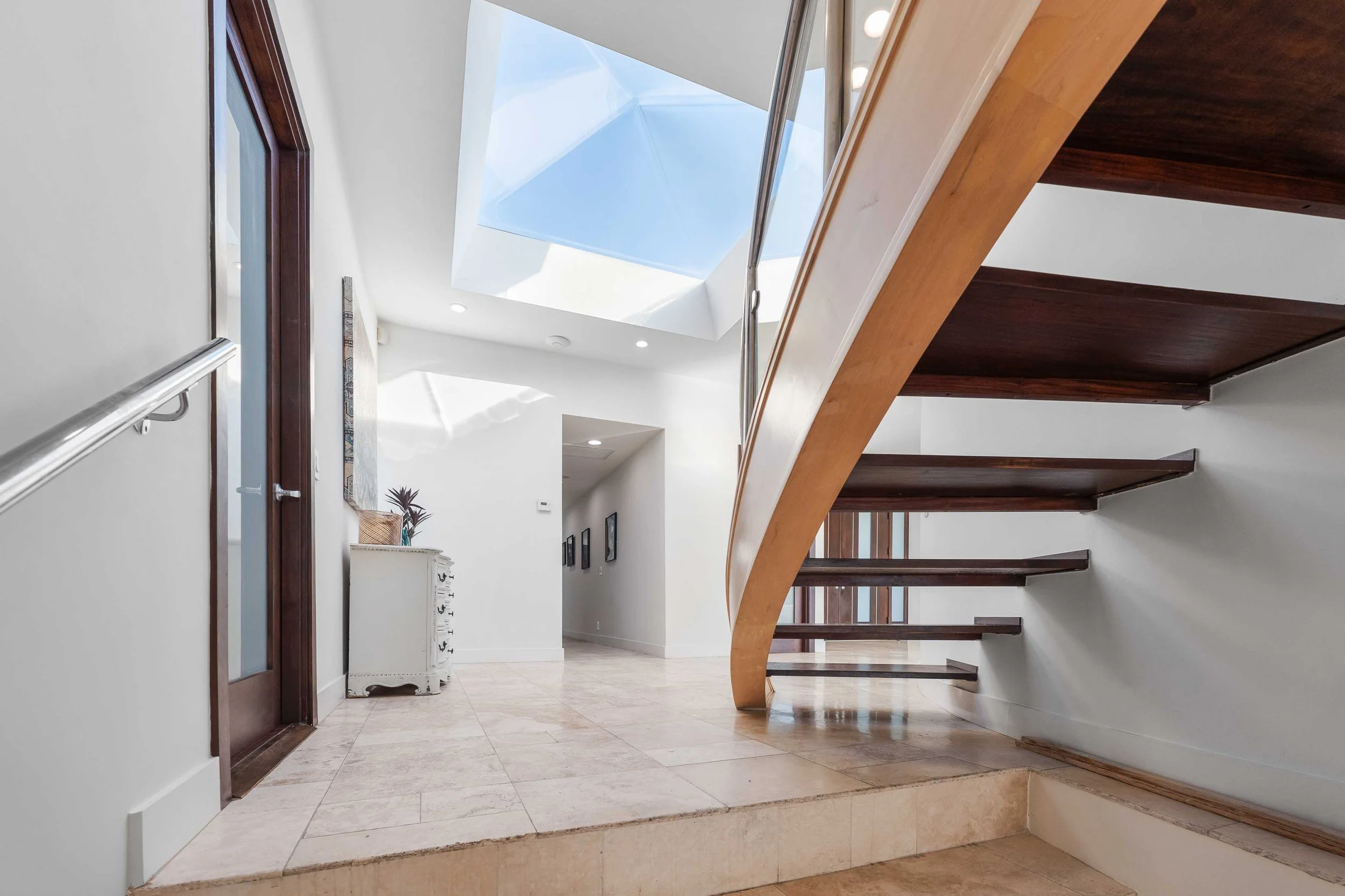 Interior view of a house with a curved staircase with dark wooden steps and a light wooden handrail, a skylight ceiling above, and a white wall with a small white console table and framed pictures in the hallway.