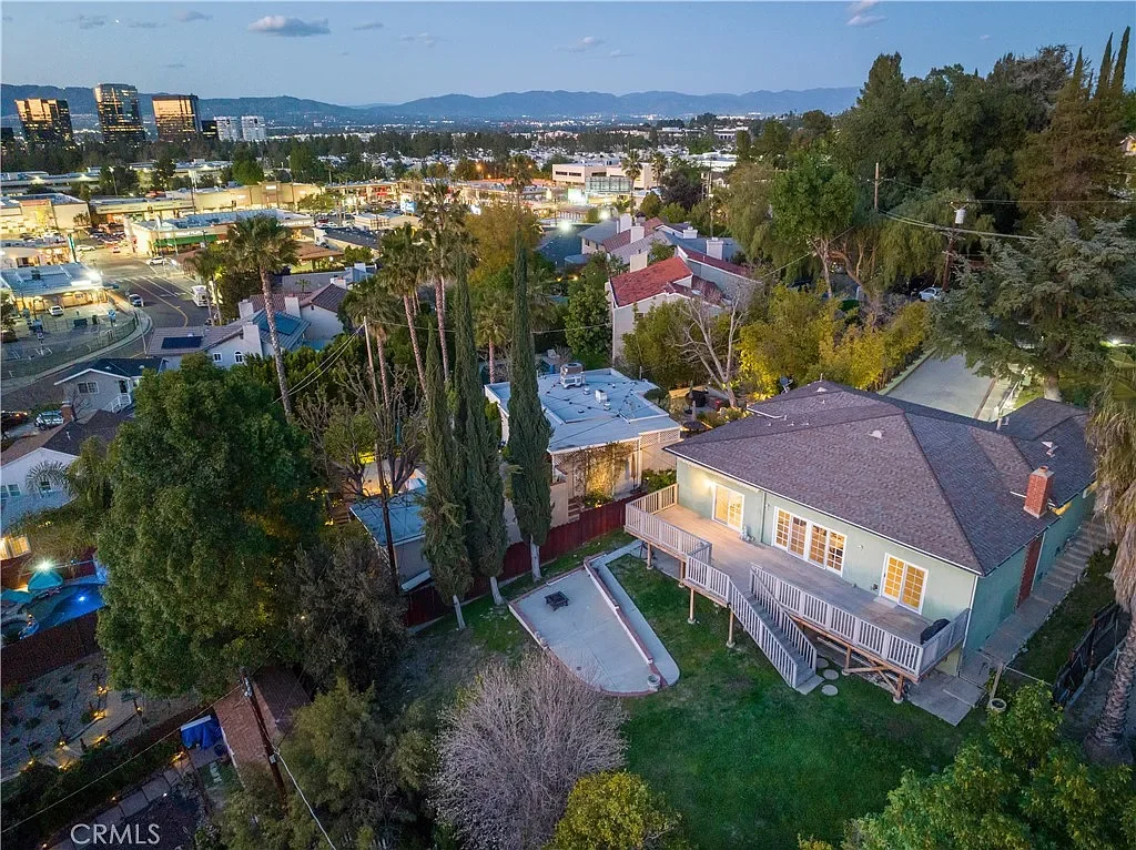 An aerial view of a residential backyard with a concrete patio, deck, and lush green lawn, surrounded by trees and neighboring houses, with a cityscape and mountains in the background at dusk.
