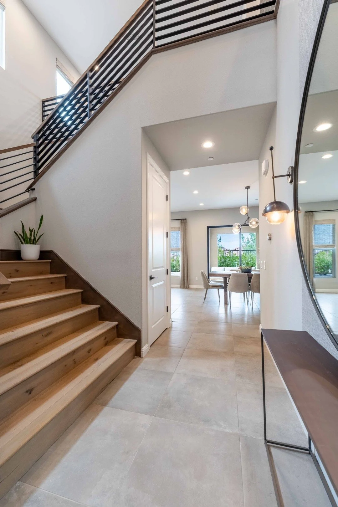 Interior view of a modern home with a staircase to the left, leading to the upper floor. The entryway has beige tiled flooring, a white door, and a wall-mounted mirror on the right. The dining area with a round table and beige chairs is visible in th