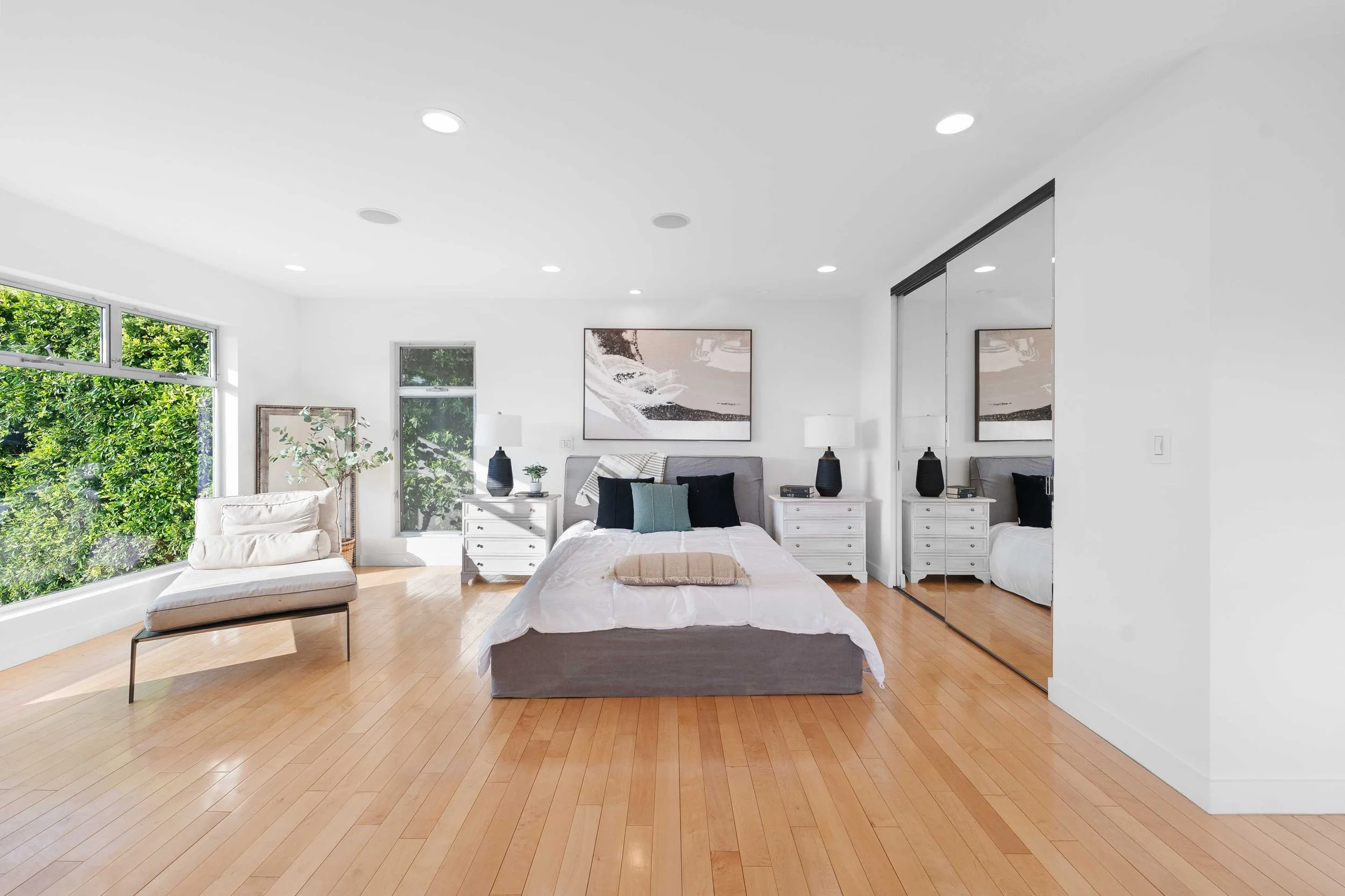 Bright modern bedroom with white walls, hardwood floors, a large bed with white bedding and black pillows, white nightstands with black lamps, large windows with green trees outside, mirrored closet doors, and a chaise lounge near the window.