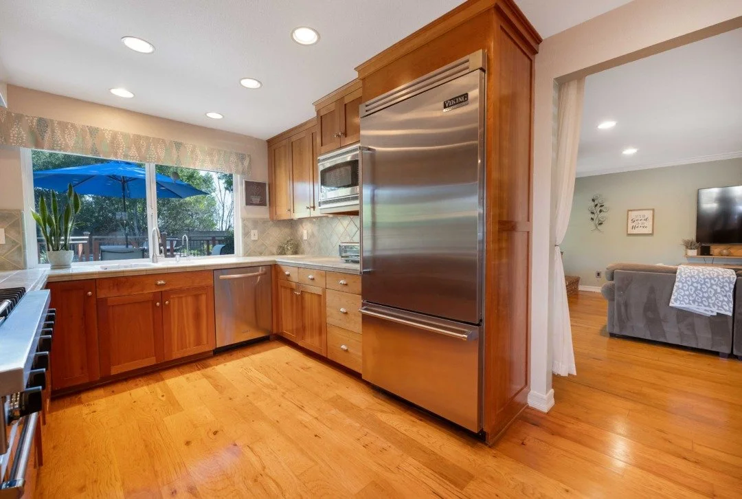 Kitchen with wooden cabinets, stainless steel appliances, window overlooking outdoor patio with blue umbrella, and hardwood floors.