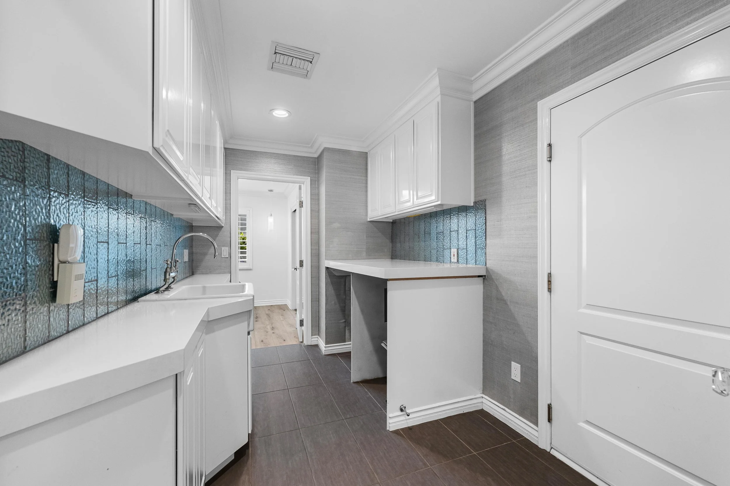 Empty laundry room with white cabinets, a laundry sink, gray textured walls, a blue tile backsplash, and dark brown floor tiles.