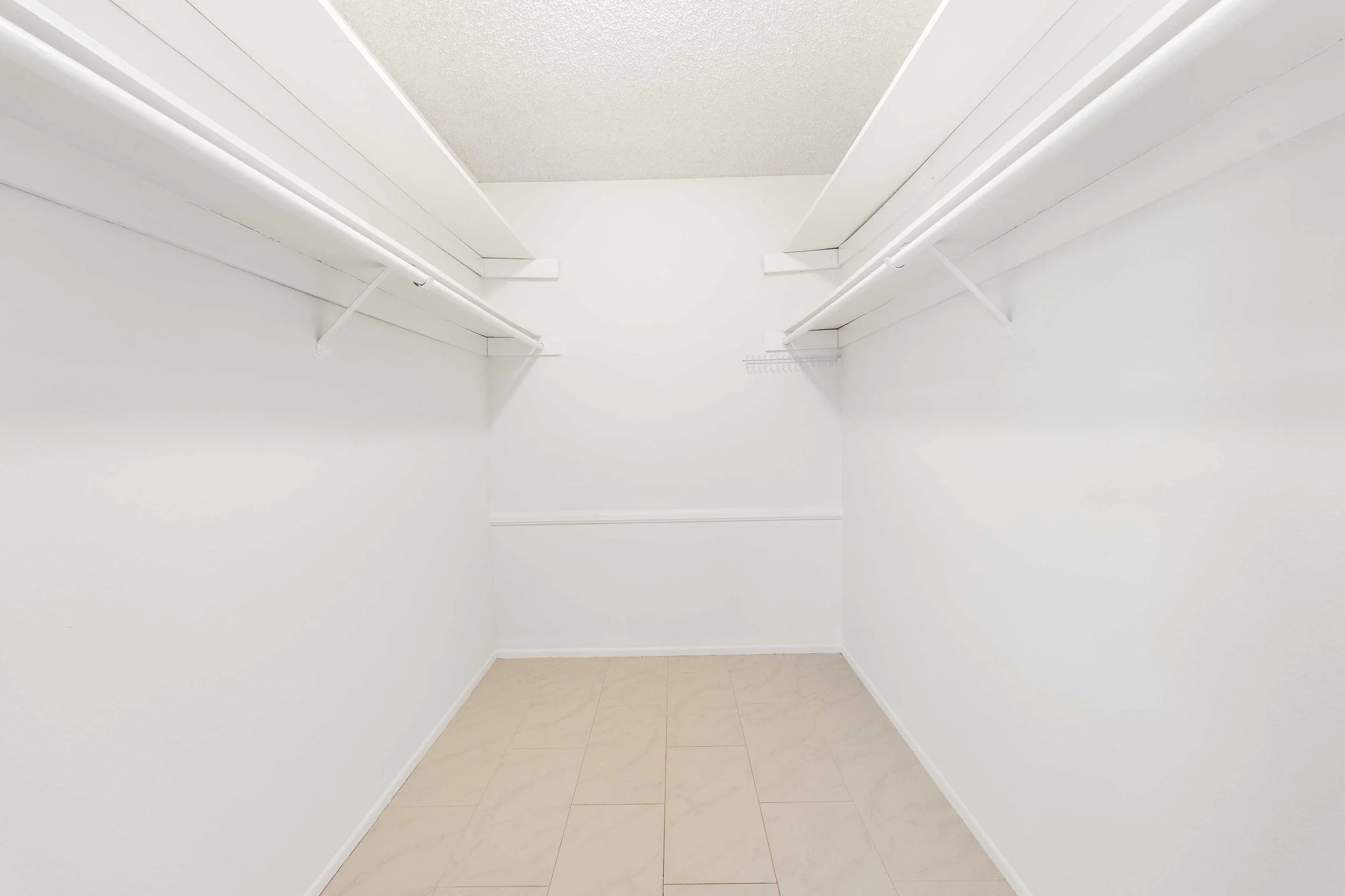 Empty white walk-in closet with beige tile floor and white shelves and hanging rods.