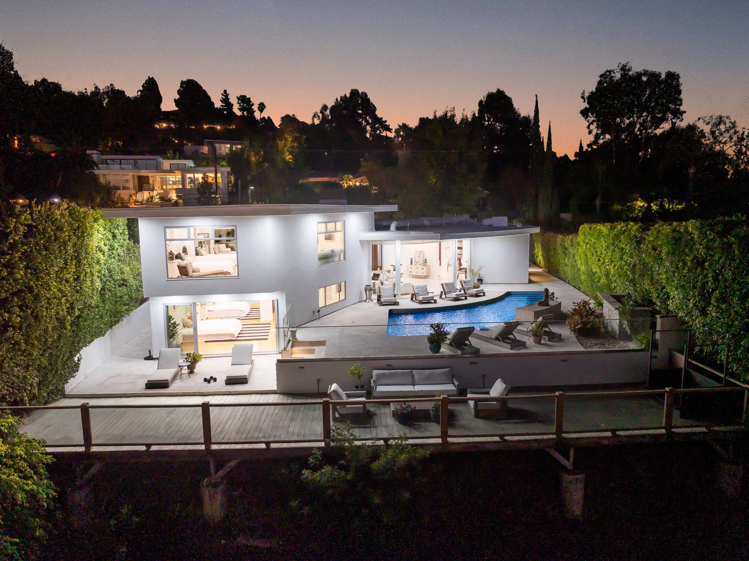 Modern two-story house with large windows, outdoor pool, lounge chairs, and patio furniture illuminated at night, surrounded by greenery and on a hillside.