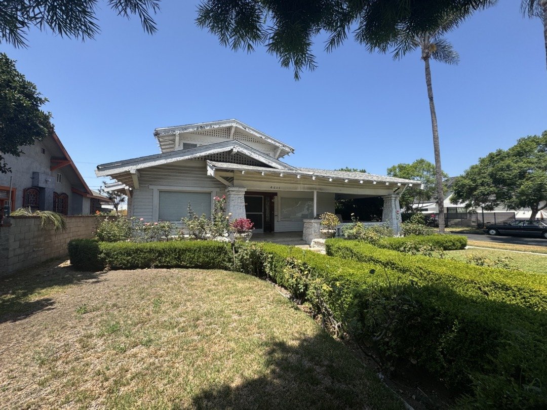 A white house with a complex roof design, front porch, surrounded by a well-maintained lawn and trimmed bushes, with a tall palm tree on the right and other trees in the background, under a clear blue sky.