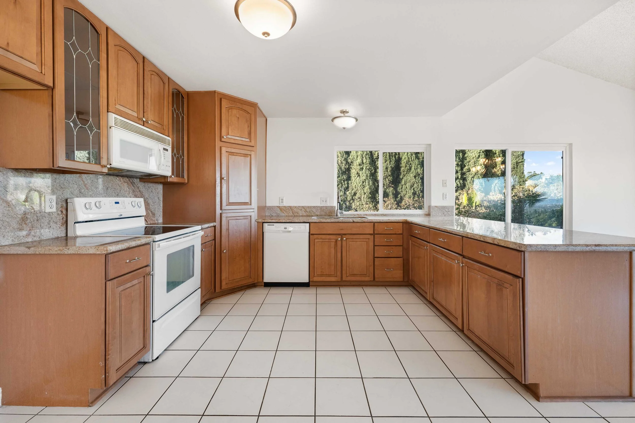 Kitchen with wooden cabinets, white appliances including stove, microwave, and dishwasher, granite countertops, white tiled floor, and windows showing greenery outside.