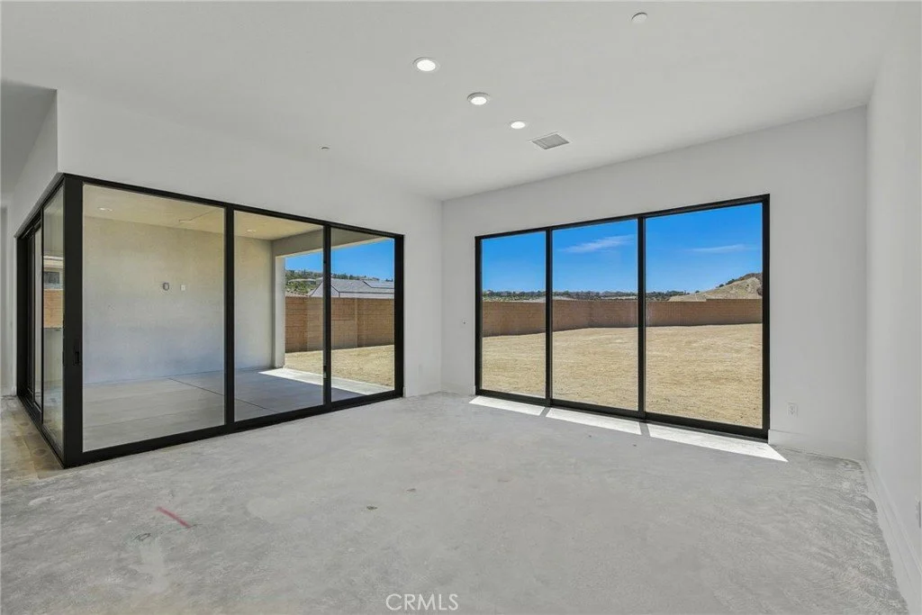 Empty living room with large black-framed sliding glass doors and windows, showing an outdoor yard and blue sky.