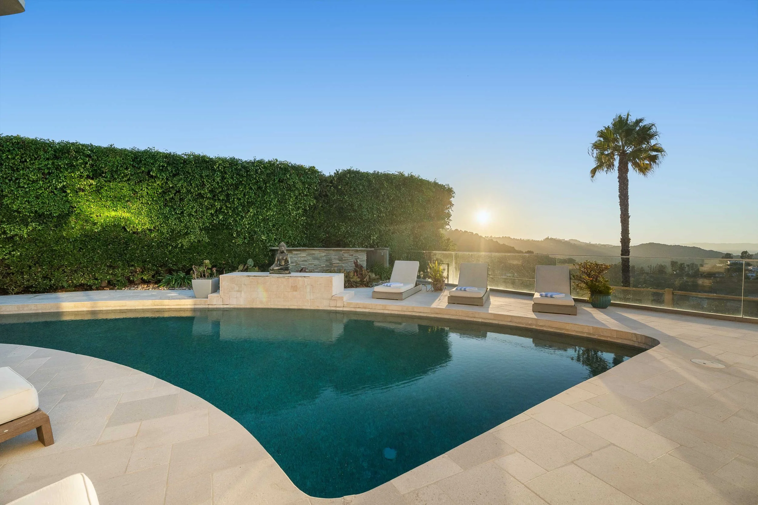 Luxury outdoor pool area with lounge chairs, plants, palm tree, and a sunset view over a hilly landscape