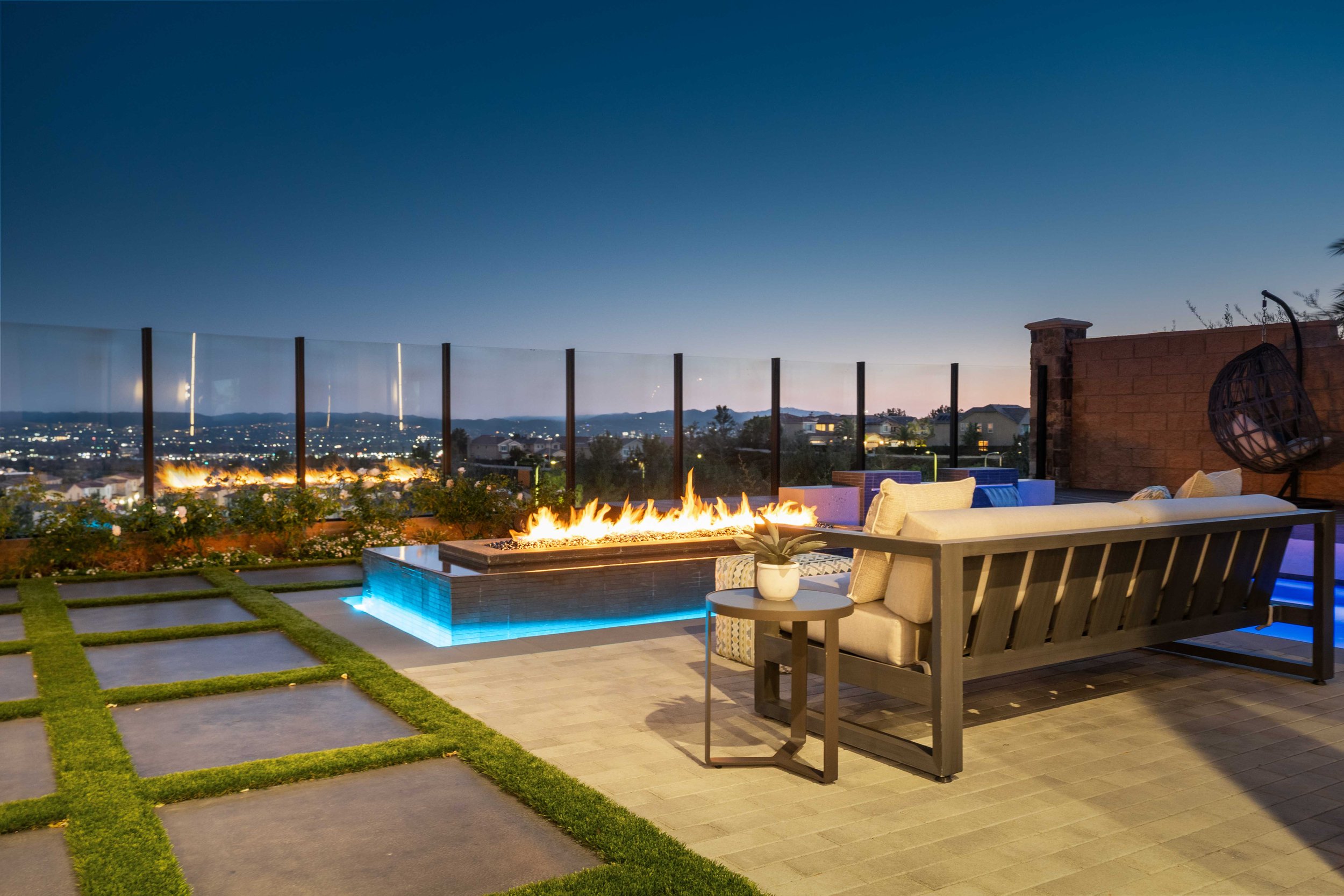 Outdoor rooftop patio at dusk with a seating area, modern fire pit, city view, and decorative lights.
