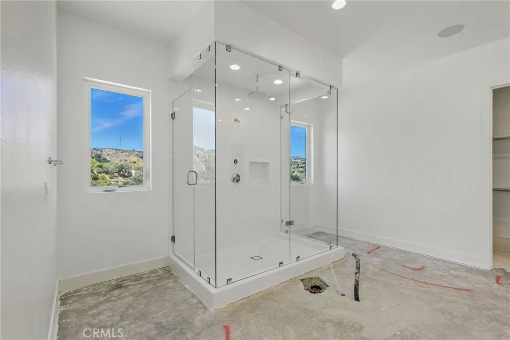 Bathroom with a glass shower enclosure, two windows showing a hilly landscape, and a concrete floor in progress.