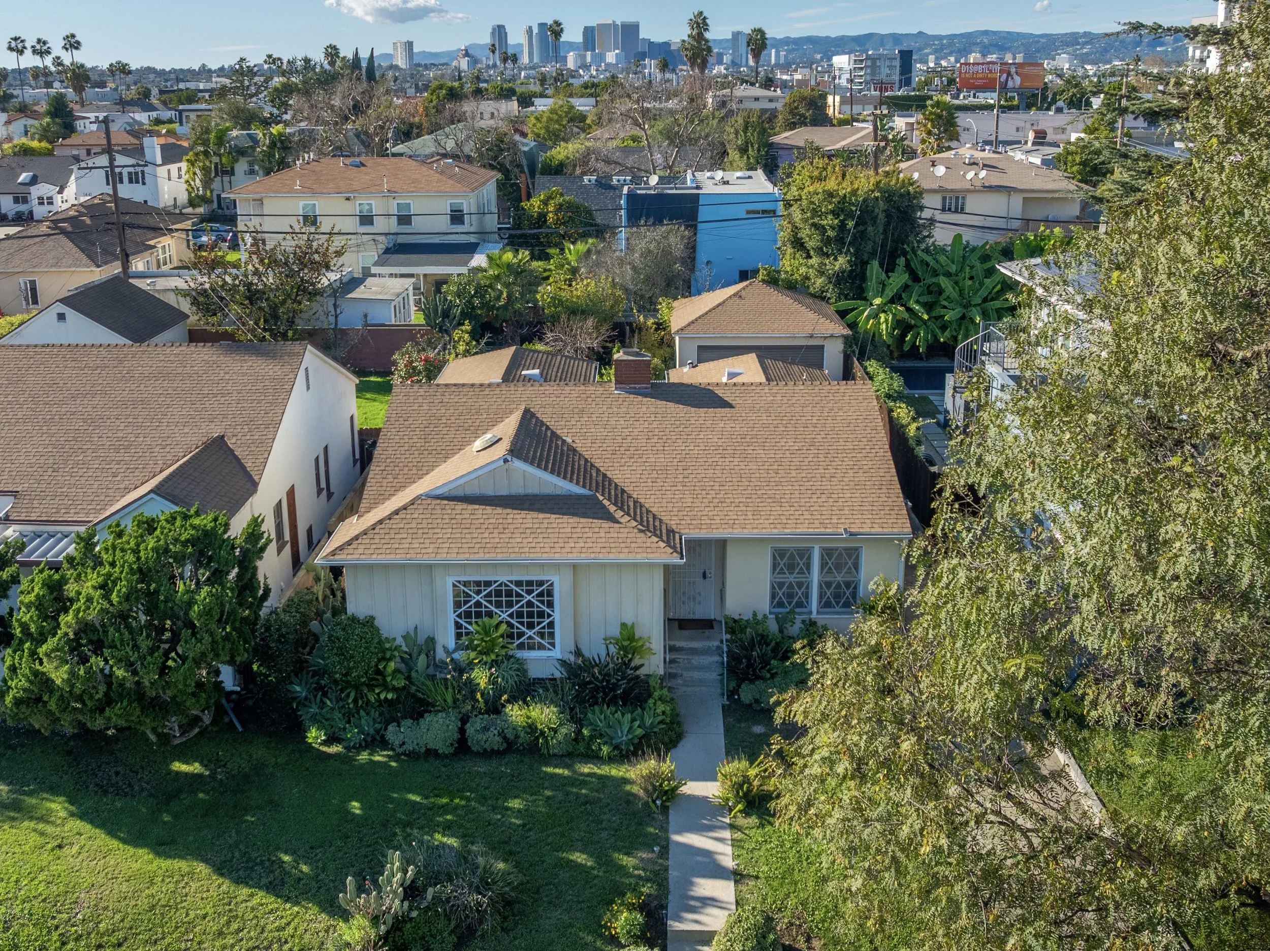A yellow single-story house with a brown shingle roof, surrounded by green plants and shrubs. There are stairs leading up to the front door, and a driveway on the right side. The front yard has a small lawn and a concrete sidewalk.