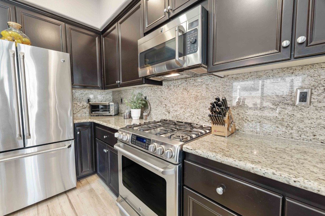Kitchen with dark cabinets, stainless steel refrigerator, microwave, oven, granite countertops, and small appliances on counter.