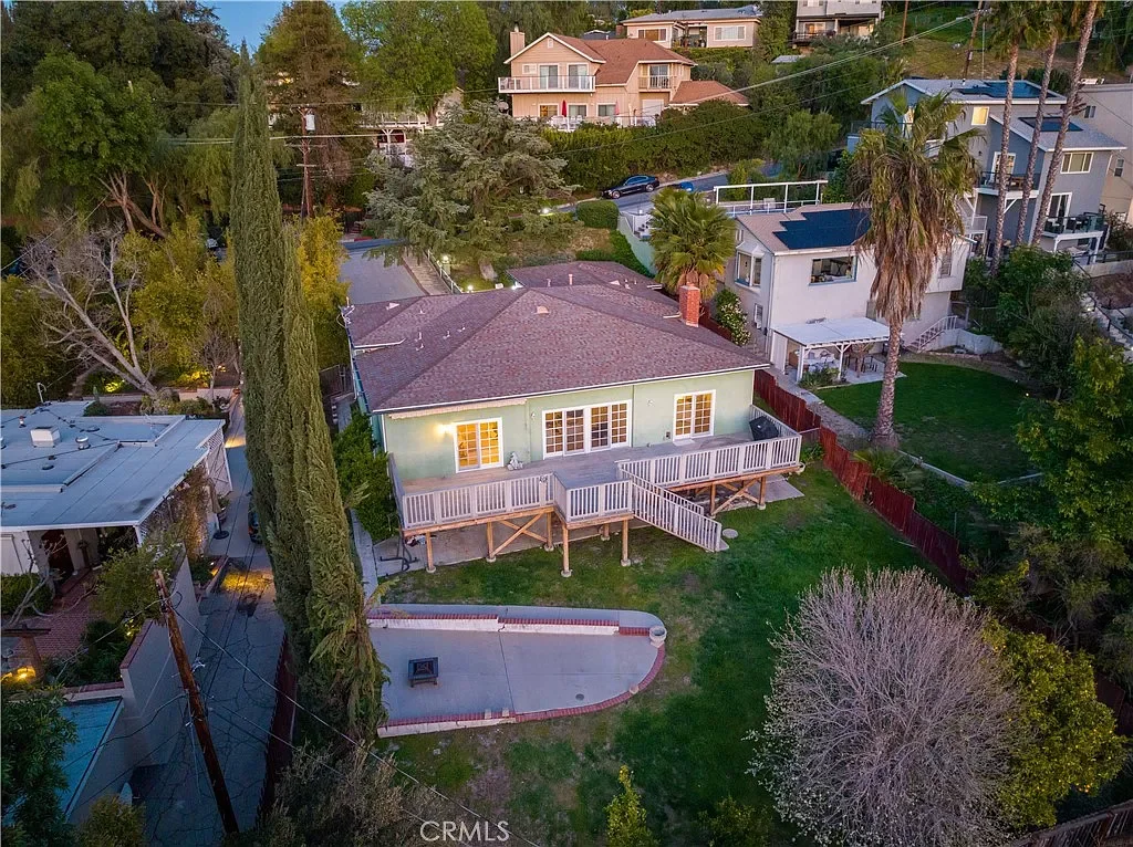 Aerial view of a backyard with a raised deck, a small concrete area, and green grass, surrounded by various trees and neighboring houses in a suburban area.