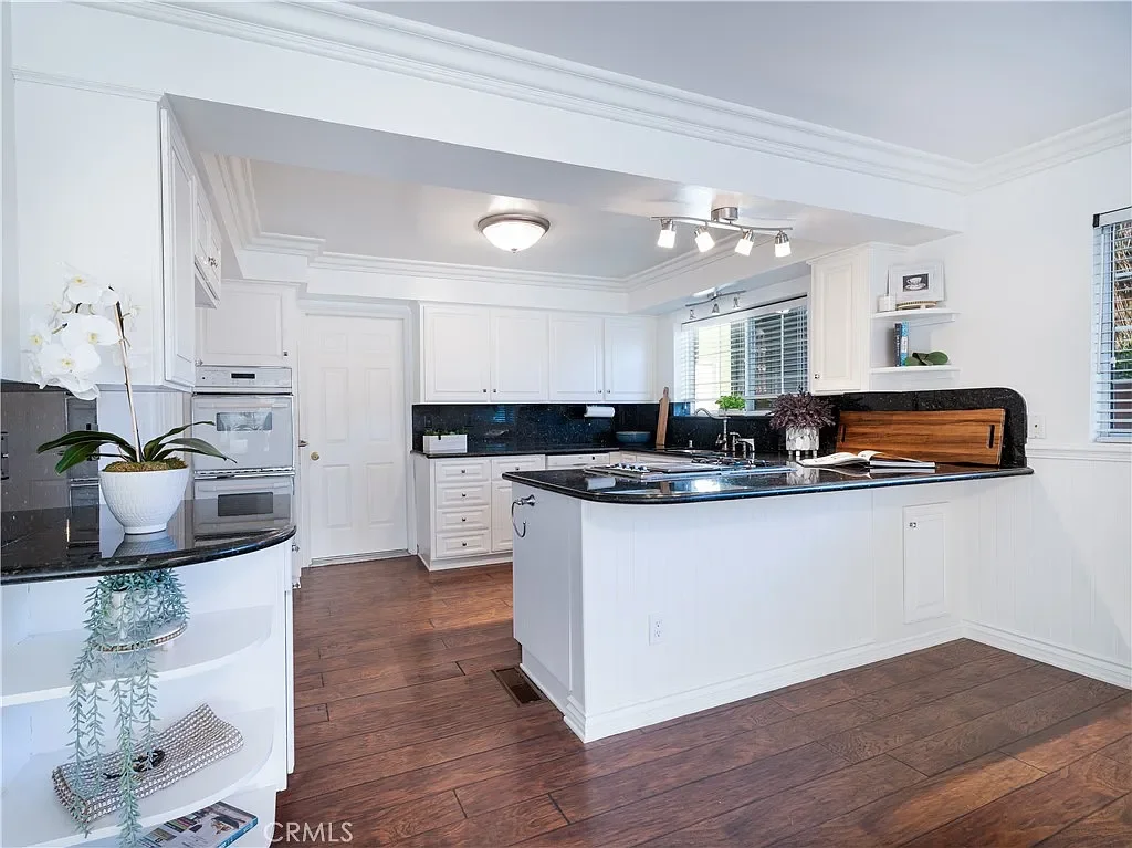 Bright white kitchen with black granite countertops, wooden flooring, and large windows with blinds.