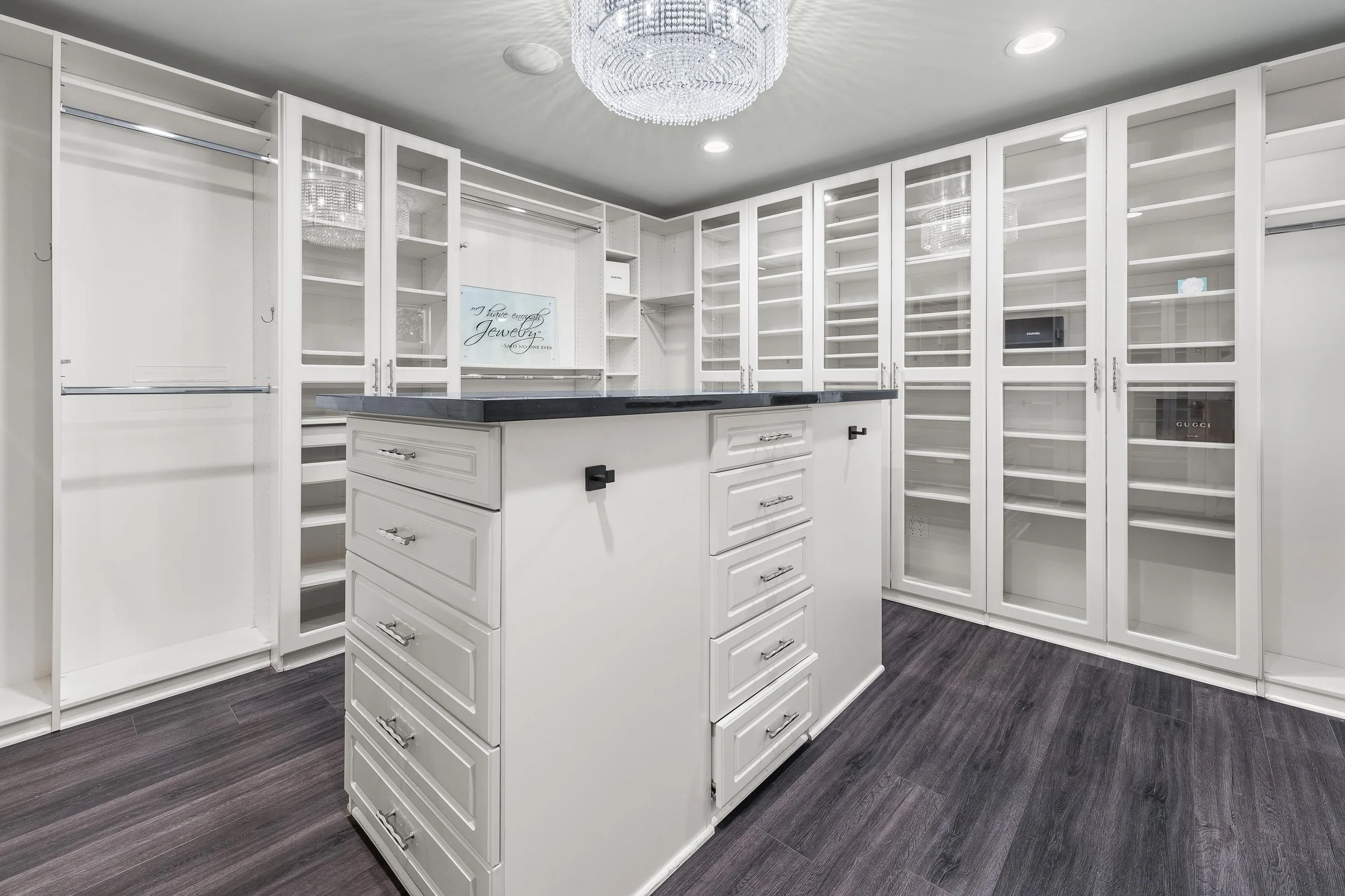 A spacious walk-in closet with white cabinetry, glass-fronted shelves, and a kitchen island with storage drawers and a dark countertop, dark wood flooring, and a chandelier hanging from the ceiling.