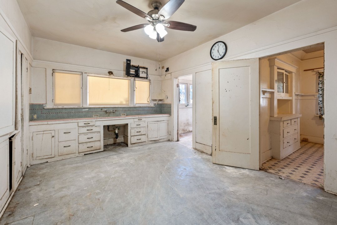 A worn, empty kitchen with white cabinets, a ceiling fan, and a clock on the wall. The countertops and floor are in poor condition.