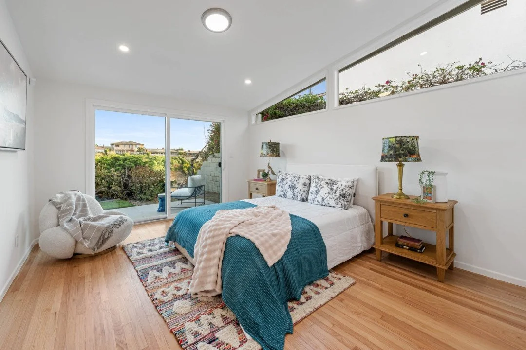 Bright bedroom with sliding glass door leading to a patio, a bed with blue bedding and floral pillows, two wooden nightstands with lamps, a cozy armchair, and a colorful area rug on light hardwood flooring. Window above the bed shows outdoor greenery