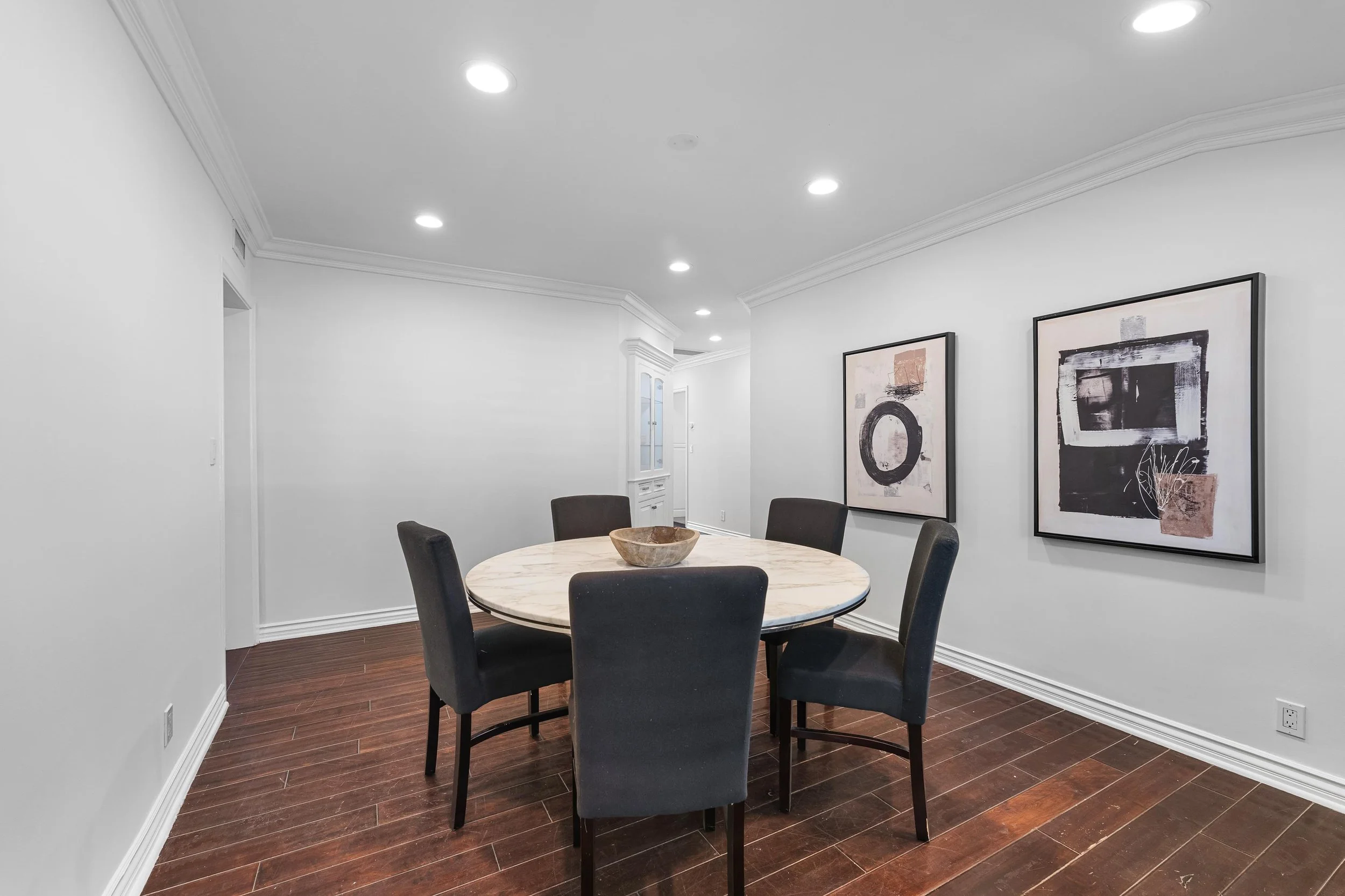 Modern dining room with a round marble table, six black chairs, and framed abstract artwork on white walls.