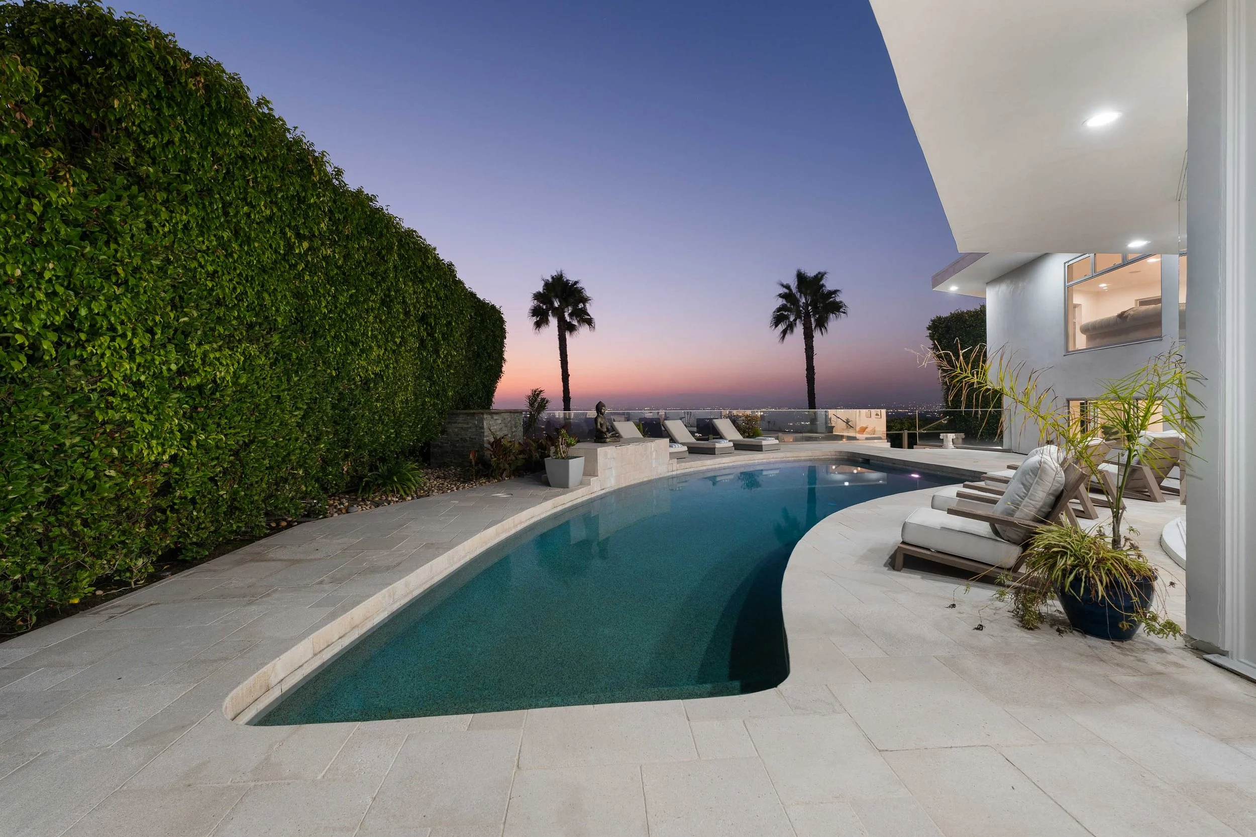 Modern backyard with a curved swimming pool, lounge chairs, potted plants, a large green hedge, and palm trees at sunset.