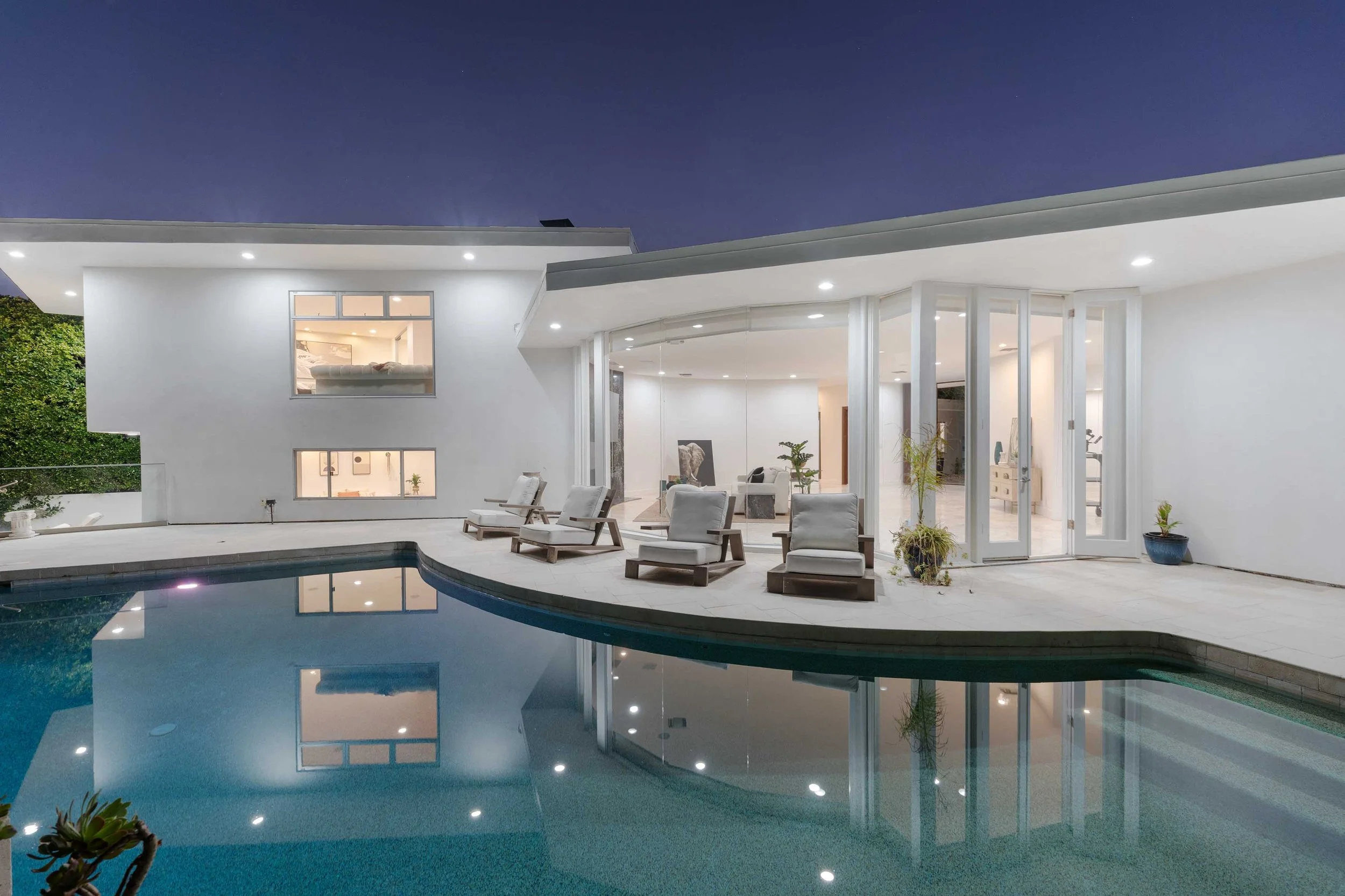 Modern house exterior at night with a swimming pool, white walls, large glass doors and windows, patio chairs, and string lights.