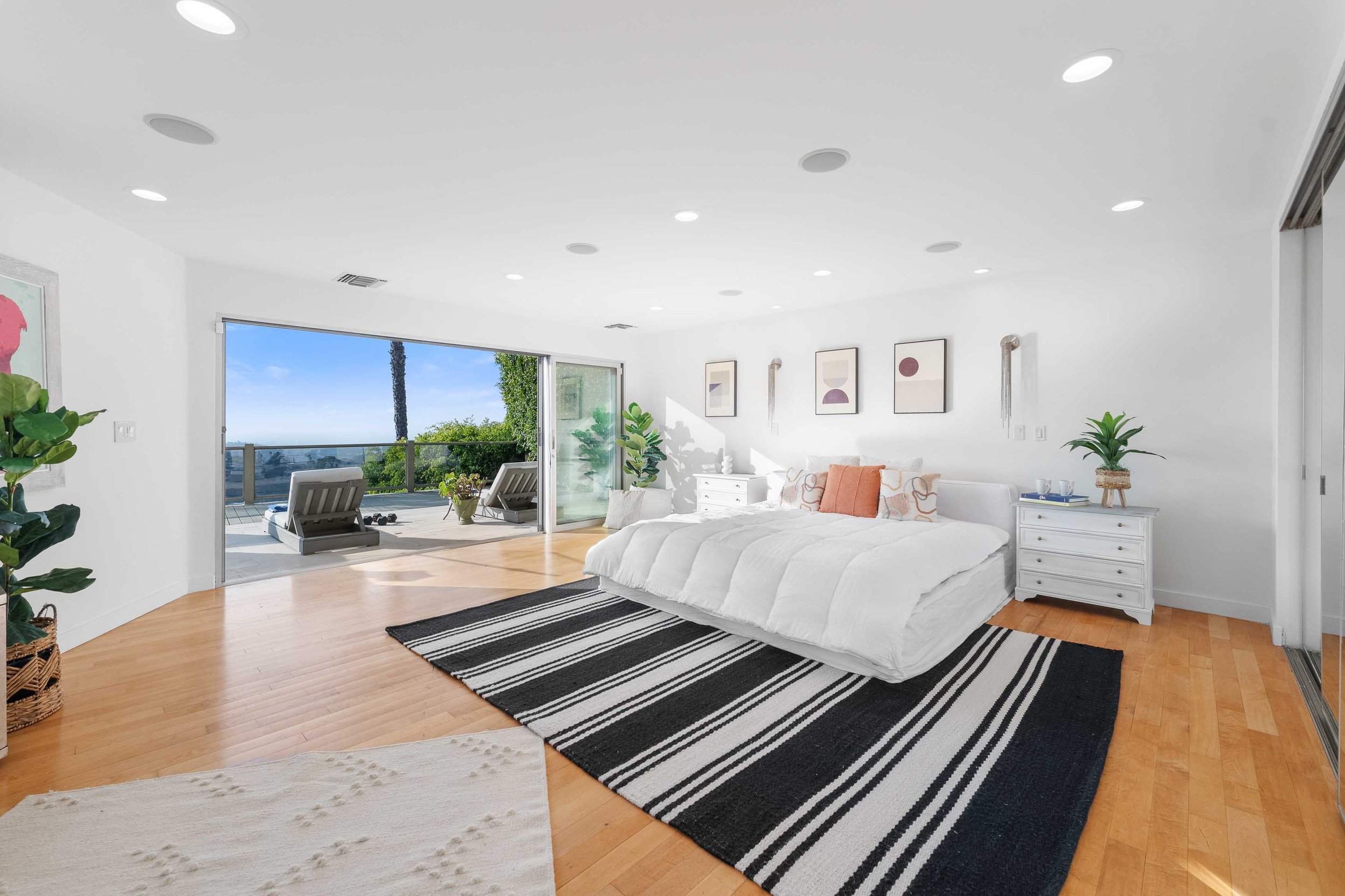 Bright bedroom with white walls, bed with white bedding and orange pillows, black and white striped rug, white nightstands, artwork on the wall, sliding glass door open to outdoor patio with lounge chairs and view of trees and sky.