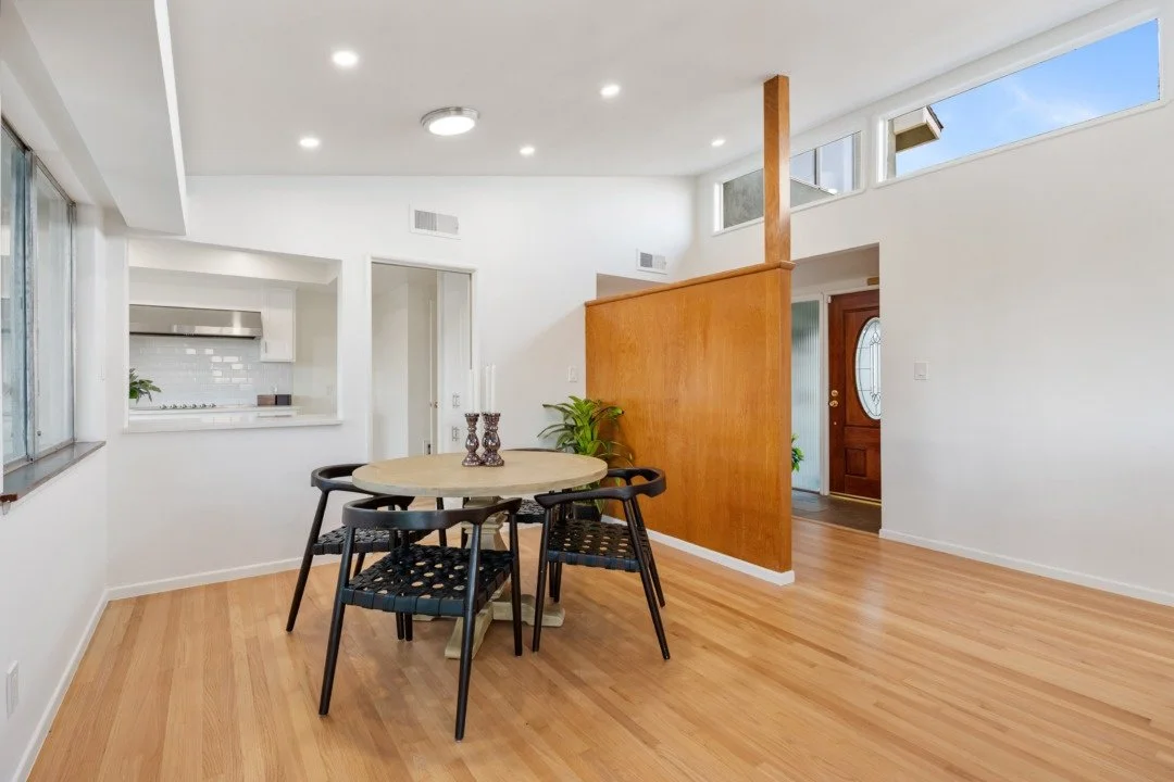 Empty dining room with round table, six black chairs, wooden floor, white walls, partial kitchen view, and front door in the background.