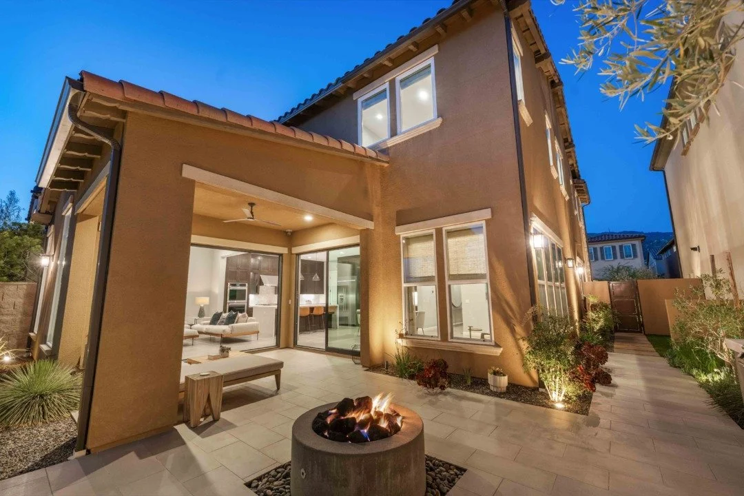 Backyard patio with outdoor fire pit, modern house with large windows, landscaped plants, and outdoor lighting at dusk.