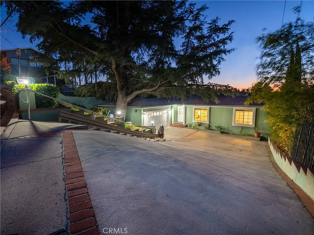 Residential house with a large driveway, green exterior walls, illuminated windows, and outdoor lighting, set amidst trees at dusk.