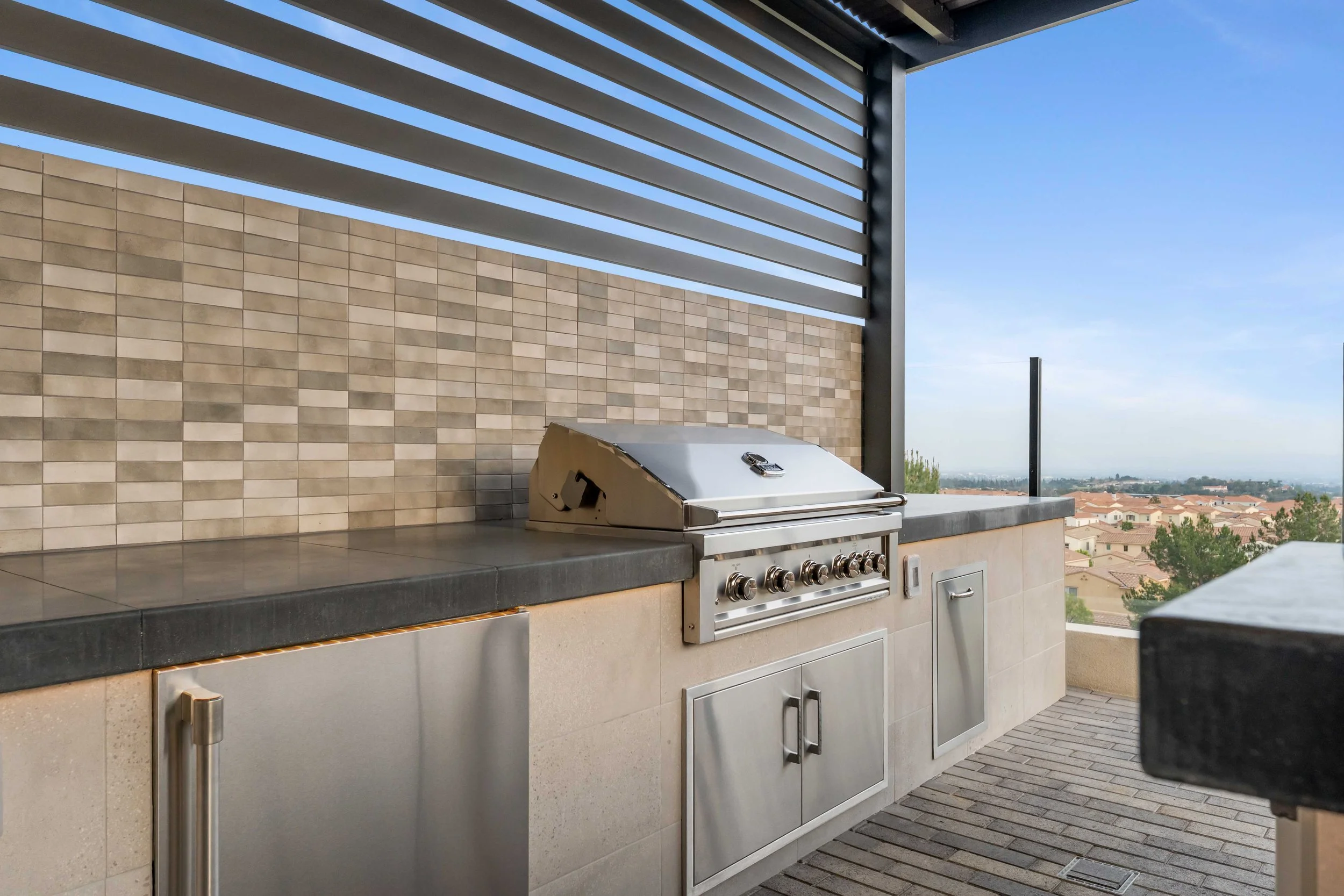 Outdoor rooftop kitchen with a stainless steel grill, black tile countertop, beige tile walls, and a railing overlooking a neighborhood with trees and houses under a clear blue sky.