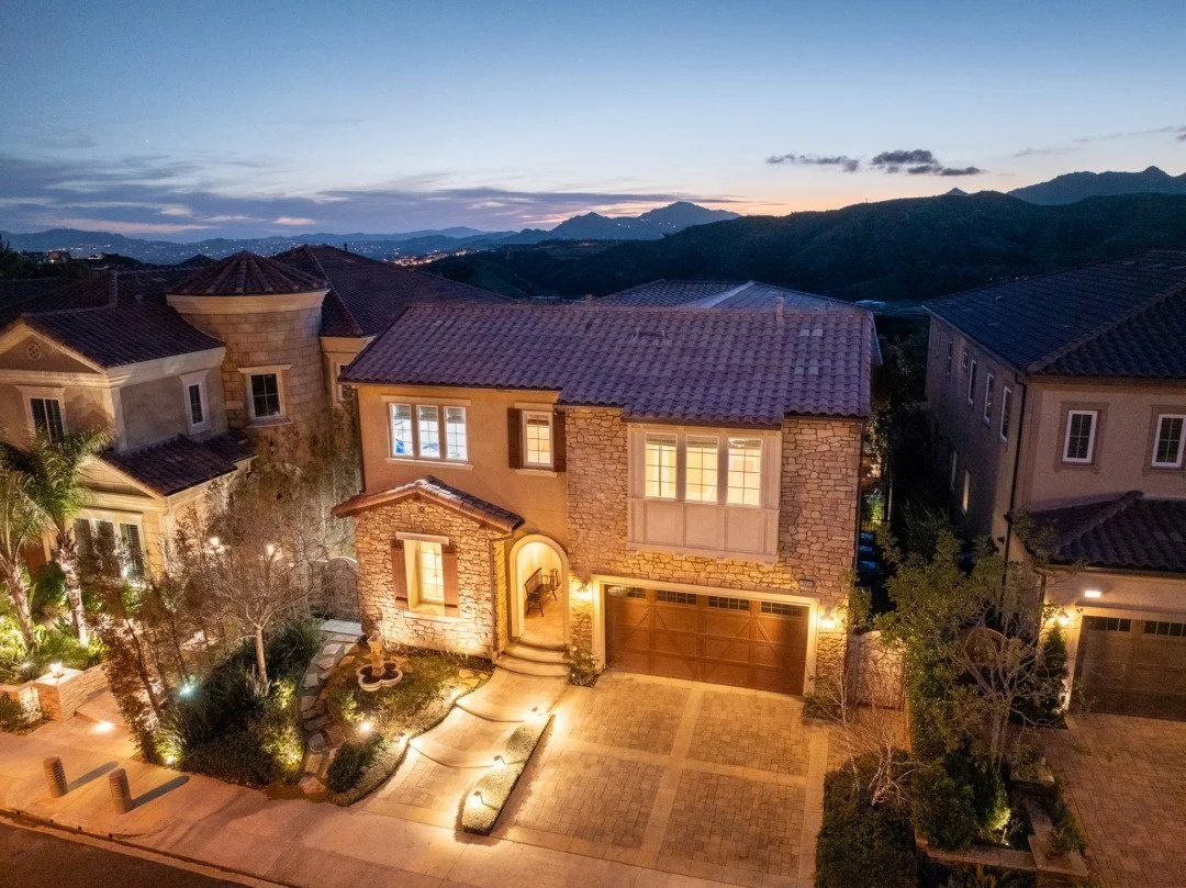 Aerial view of a house with a front yard at dusk, neighboring homes, mountains in the background, and city lights in the distance.