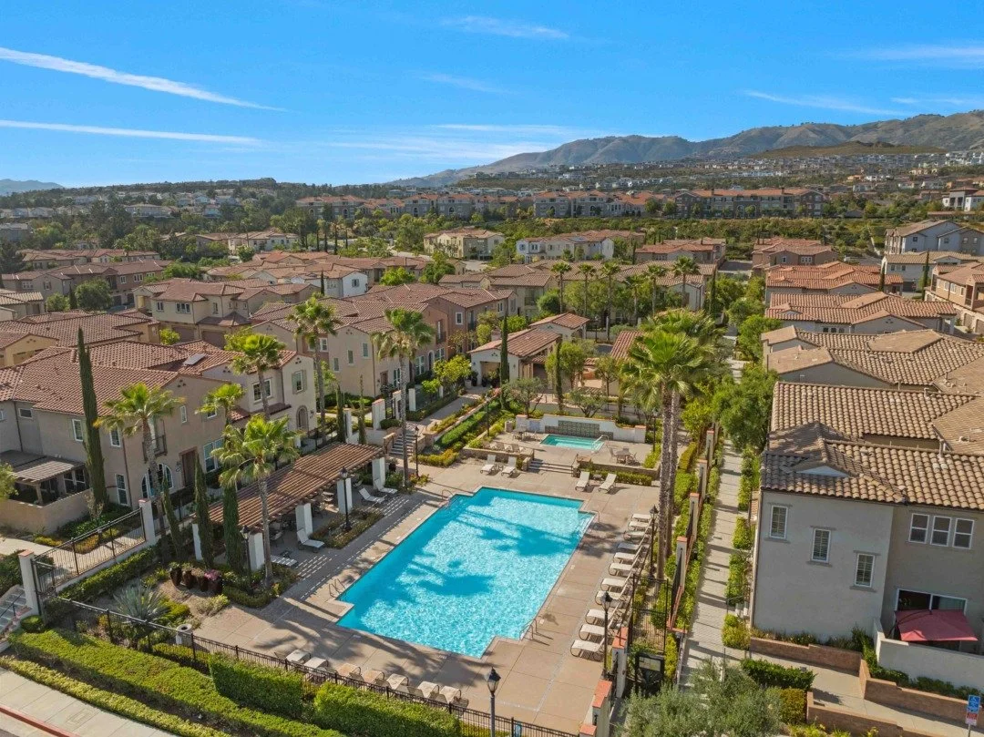 Aerial view of a residential community with swimming pools, palm trees, and beige houses with red tile roofs, set against hills under a blue sky.