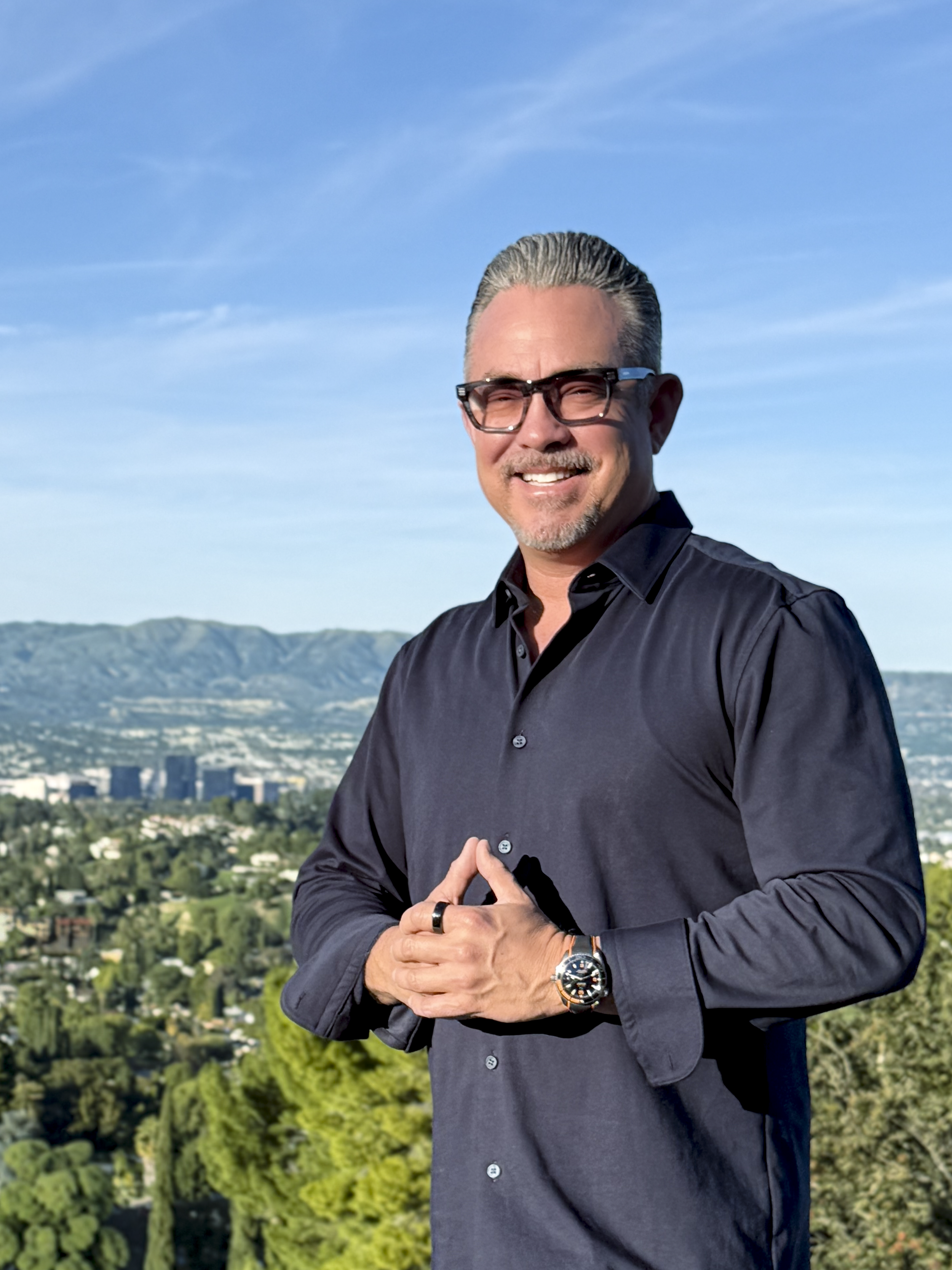 Real estate agent and broker Collin St. Johns' profile image on top of a hill wearing a blue button down shirt, watch, and glasses. The Warner Center can be seen behind him in the background.