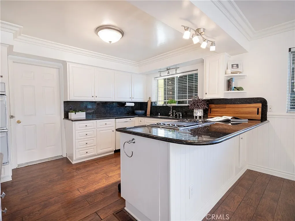 White kitchen with dark granite countertops, wooden flooring, and multiple cabinets. There's a window above the sink, a small shelf with books and decor, and a kitchen island with a stove on top.