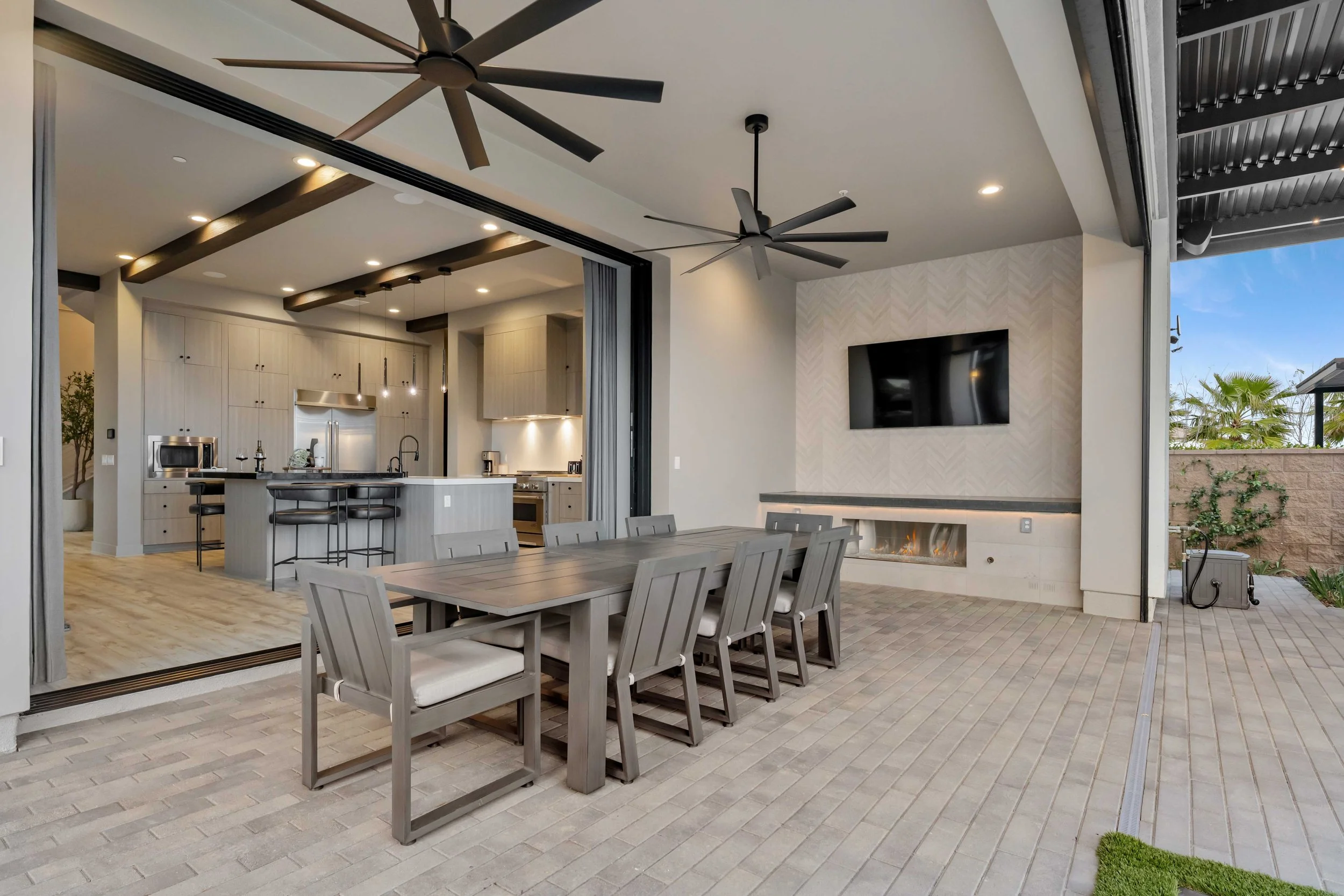 Modern outdoor patio with a dining table and chairs, a fireplace, and a view of the open kitchen and living space indoors.