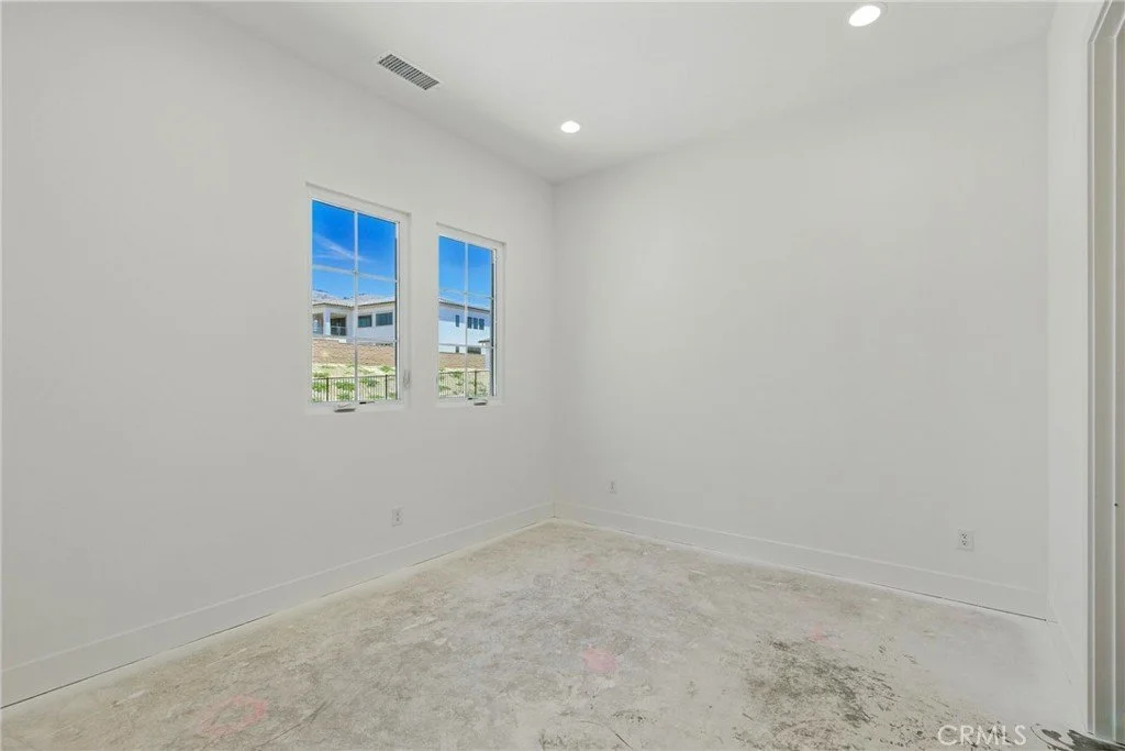 Empty white room with two windows, partially carpeted floor, and a ceiling vent, natural light coming in.