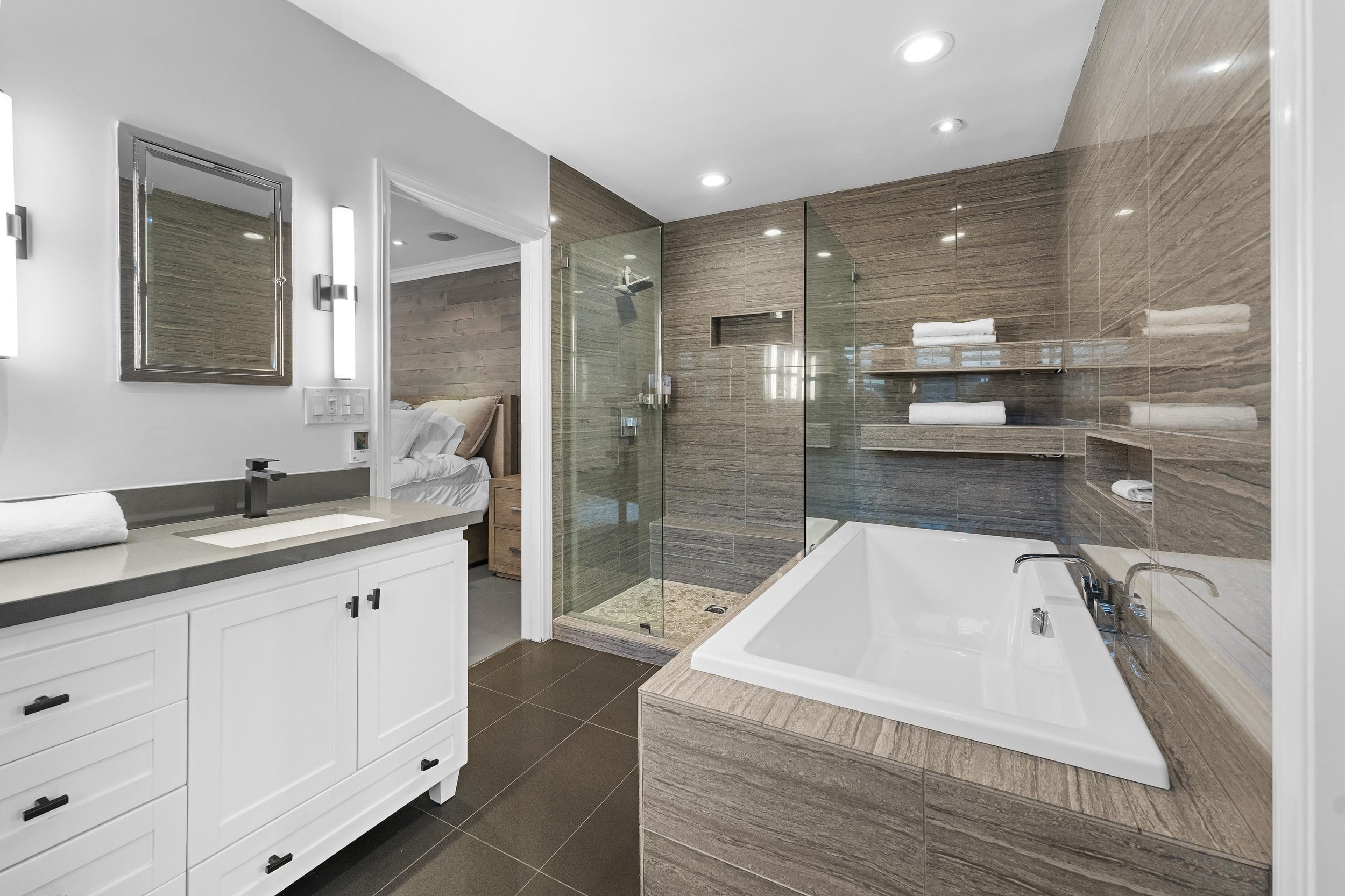 Modern bathroom with a glass shower, bathtub, white vanity, and towels, adjacent to a bedroom.