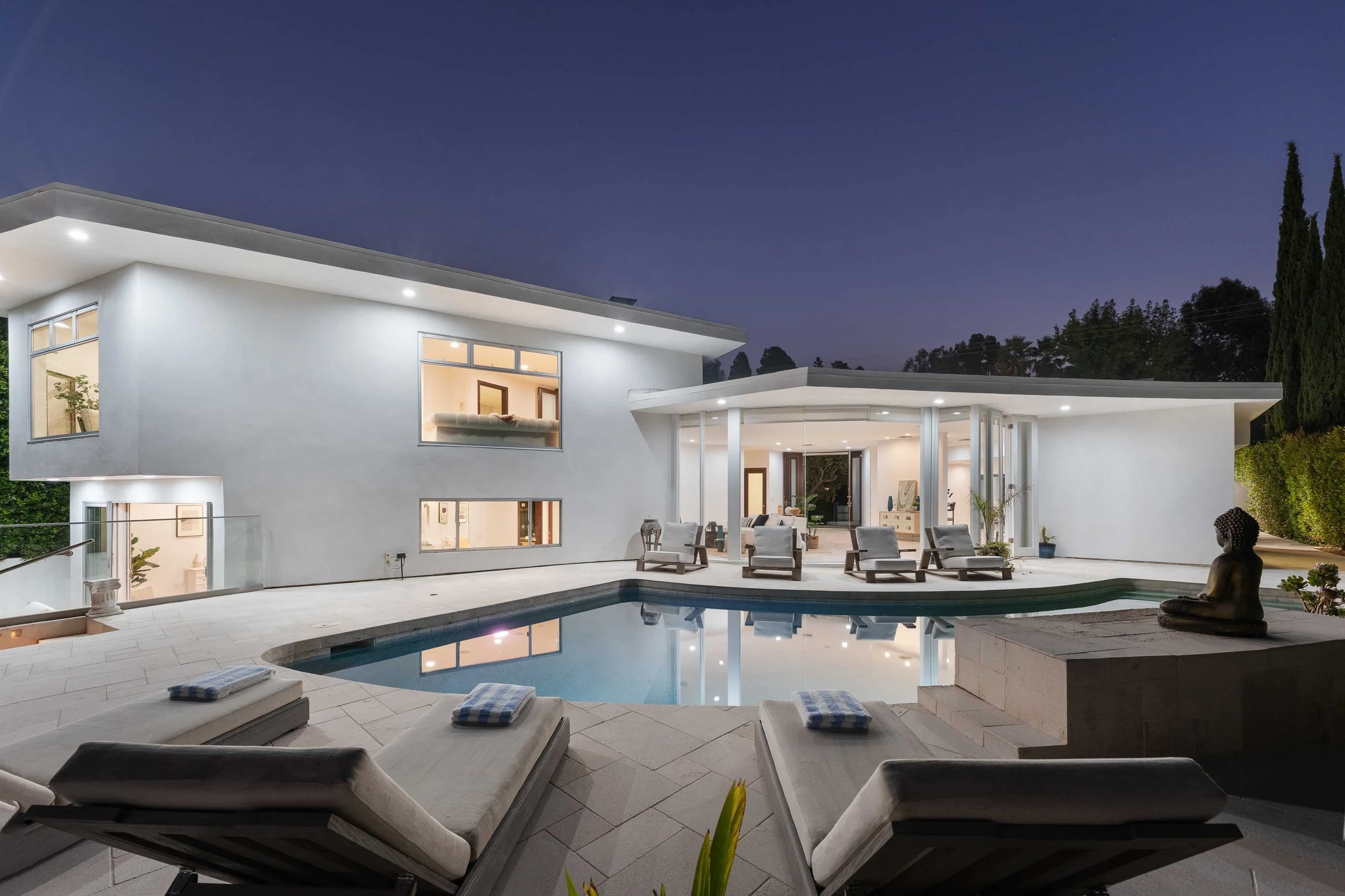 Modern white house with large windows, swimming pool, and outdoor lounge area at night