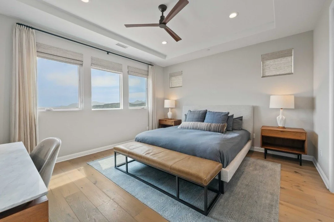 A modern bedroom with a large bed, three side tables with lamps, a bench at the foot of the bed, large windows with curtains, a ceiling fan, and a view of mountains outside.