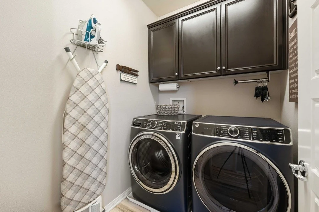 Laundry room with a washing machine and dryer, dark cabinets, wall-mounted iron, ironing board, and various small storage items.