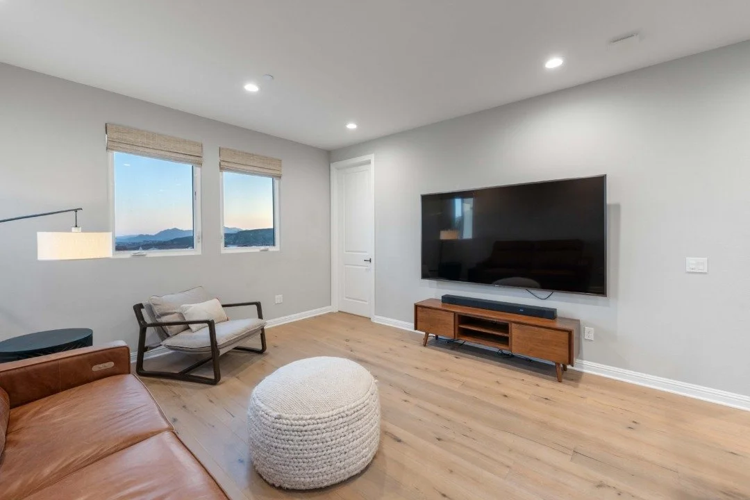 A modern living room with a wall-mounted flat-screen TV, two windows with blinds showing mountains outside, a brown leather sofa, a white knitted pouf, a black armchair with a cushion, a wooden TV stand, and a door.